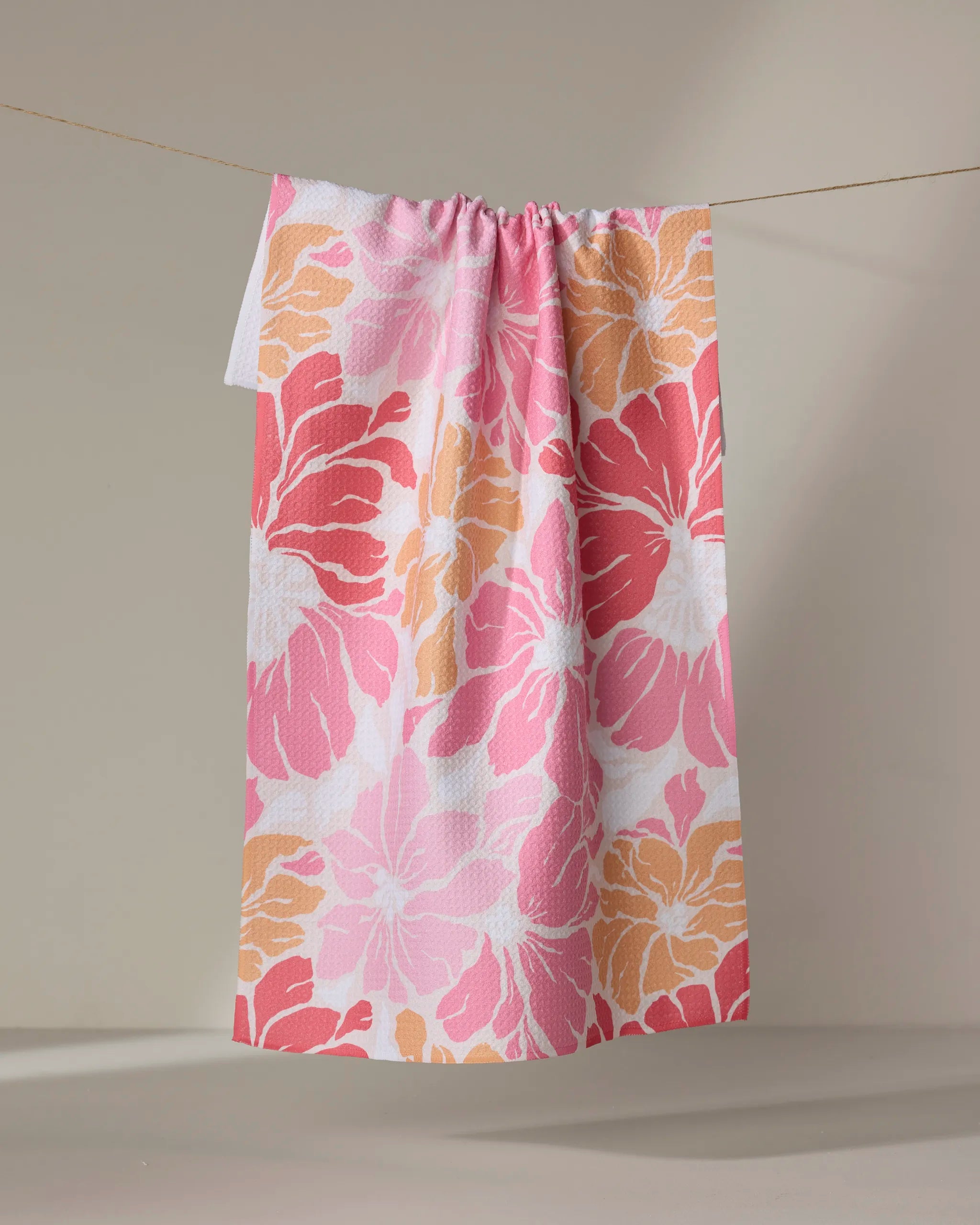 The Sunset Floral tea towel by Marina Solodka, featuring a bold pink, peach, and white floral design, dries quickly as it hangs against a neutral backdrop.