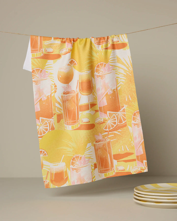 The "Sunset Happy Hour" tea towel by Geometry showcases orange and yellow tropical patterns with illustrated iced drinks, lemon slices, and ice cubes, hanging from a string against a neutral background. Eco-friendly and vibrant!.