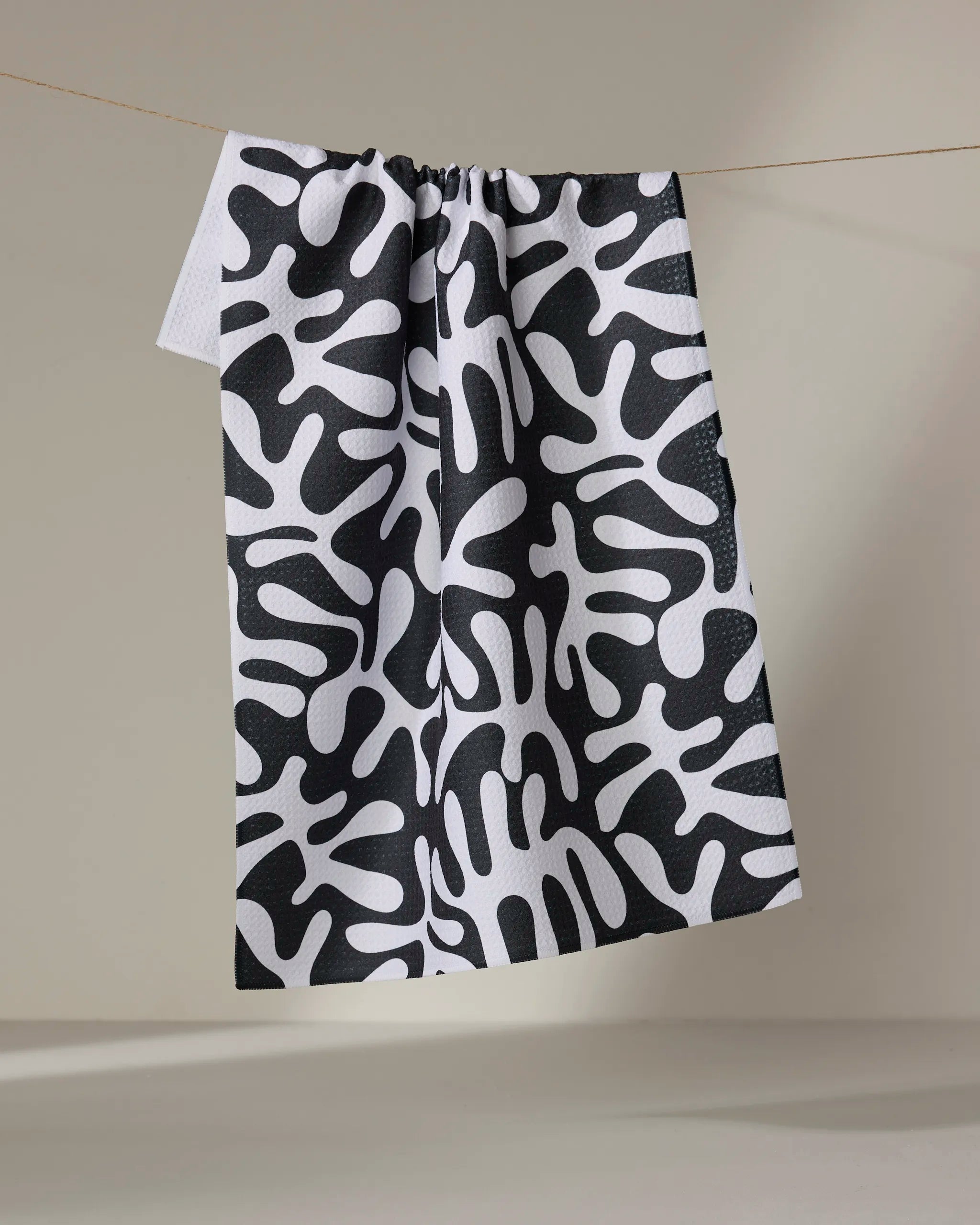 Sunset" by Marina Solodka, a recycled kitchen towel featuring a bold black and white abstract pattern of organic shapes, hangs on a line against a plain beige background.