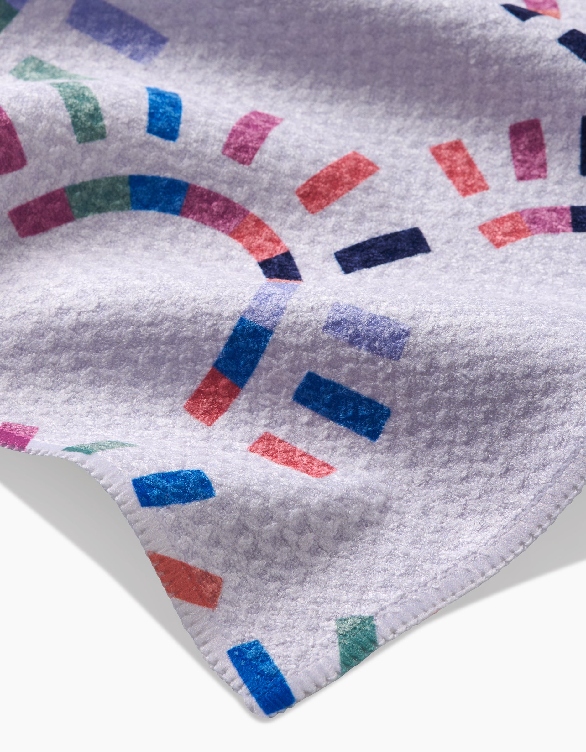 A close-up of Claire Iglesias Sunshine Rainbow towel, made from ultra-absorbent Geoweave fabric with a colorful geometric pattern in blue, purple, green, red, and pink. The white towel features neatly stitched edges.