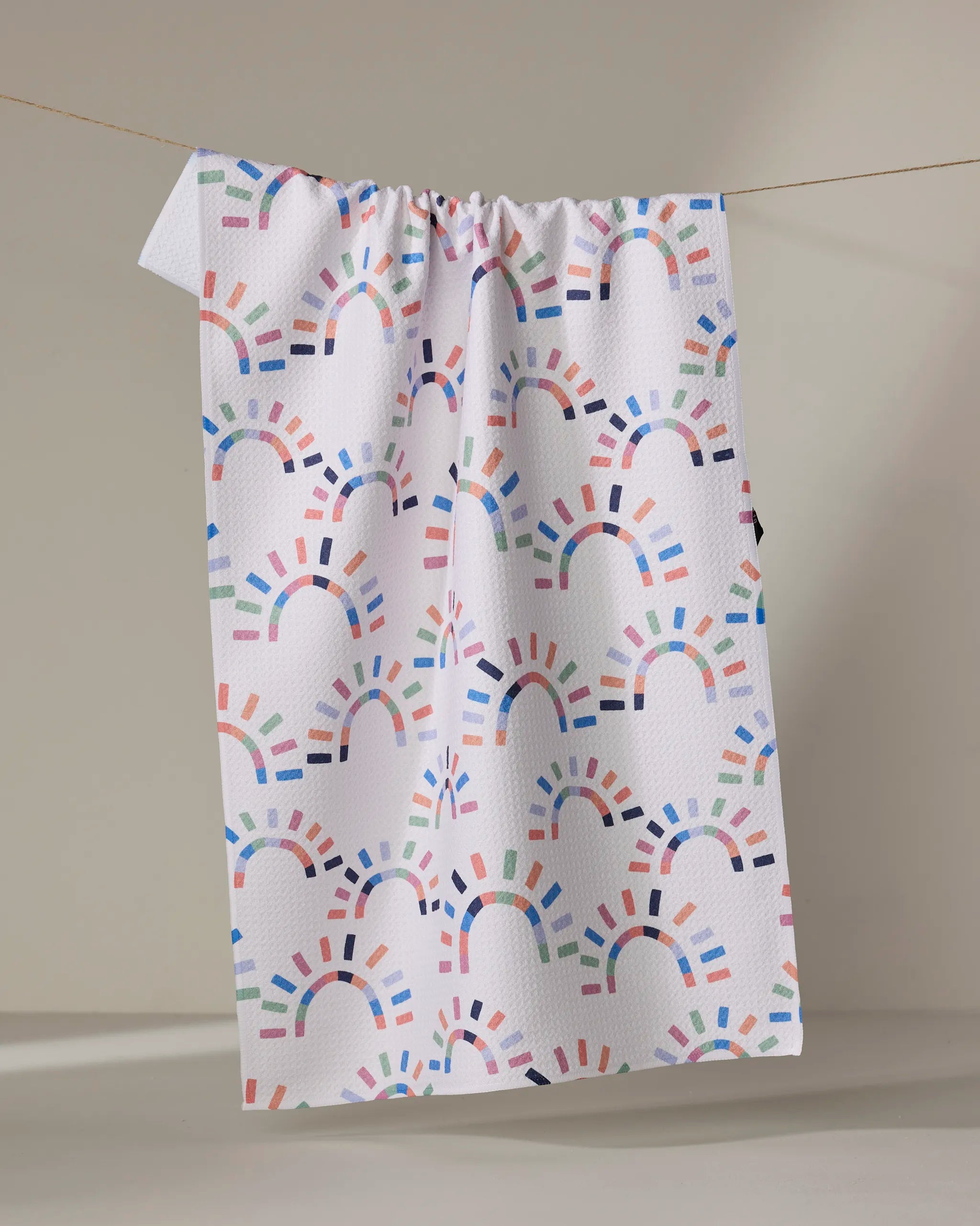 The "Sunshine Rainbow" tea towel by Claire Iglesias features colorful, semi-circular rainbow patterns and hangs on a line against a neutral background.