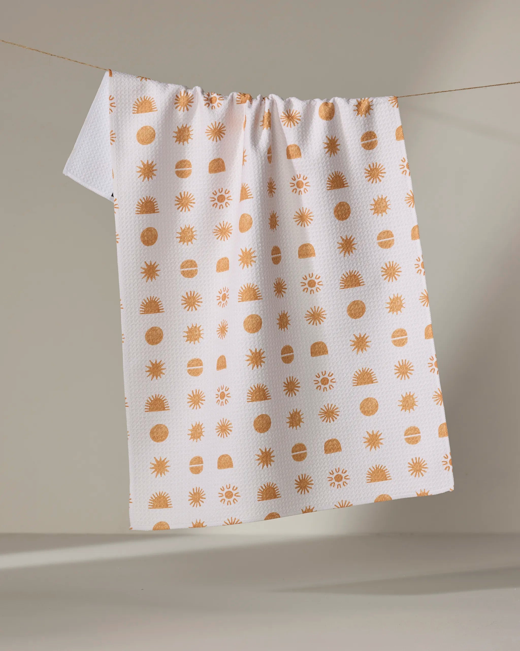 The Jen B. Peters "Sunshine" white tea towel hangs on a line, showcasing its textured surface and simple repeating orange sun, dot, and semi-circle pattern against a softly lit plain background.
