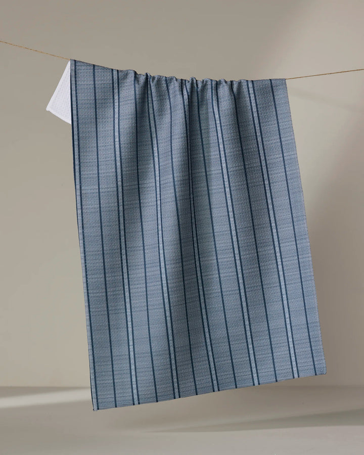 The Buy Guide’s Super Stripe - Navy kitchen towel, made from recycled materials, hangs from a thin line with blue and gray vertical stripes and a subtle texture—perfect as an absorbent dish towel against a neutral beige background.