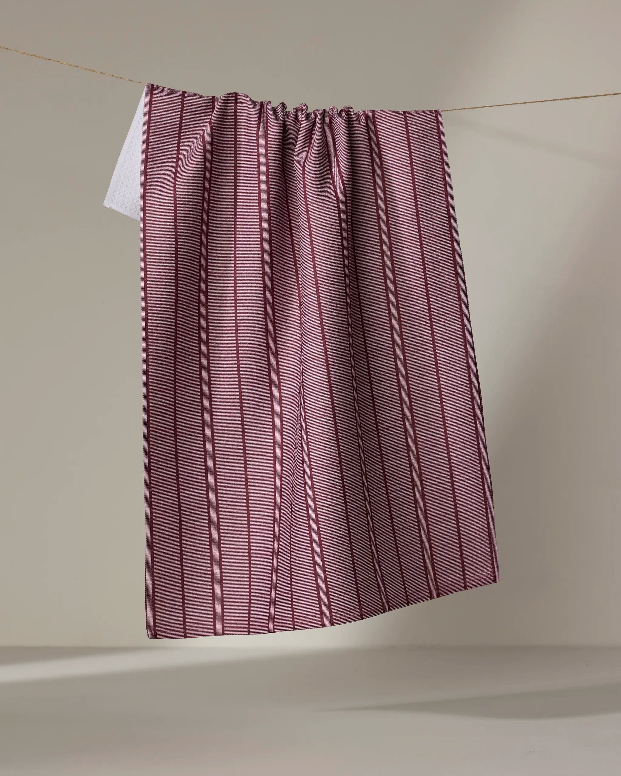The Super Stripe - Wine kitchen towel by The Buy Guide, featuring red and white stripes, hangs neatly over a thin clothesline against a plain beige background.
