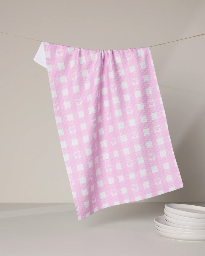 The Geometry Super Sweet Check kitchen towel, featuring pink and white checks with heart patterns, hangs on a clothesline above stacked white plates against a light gray background.