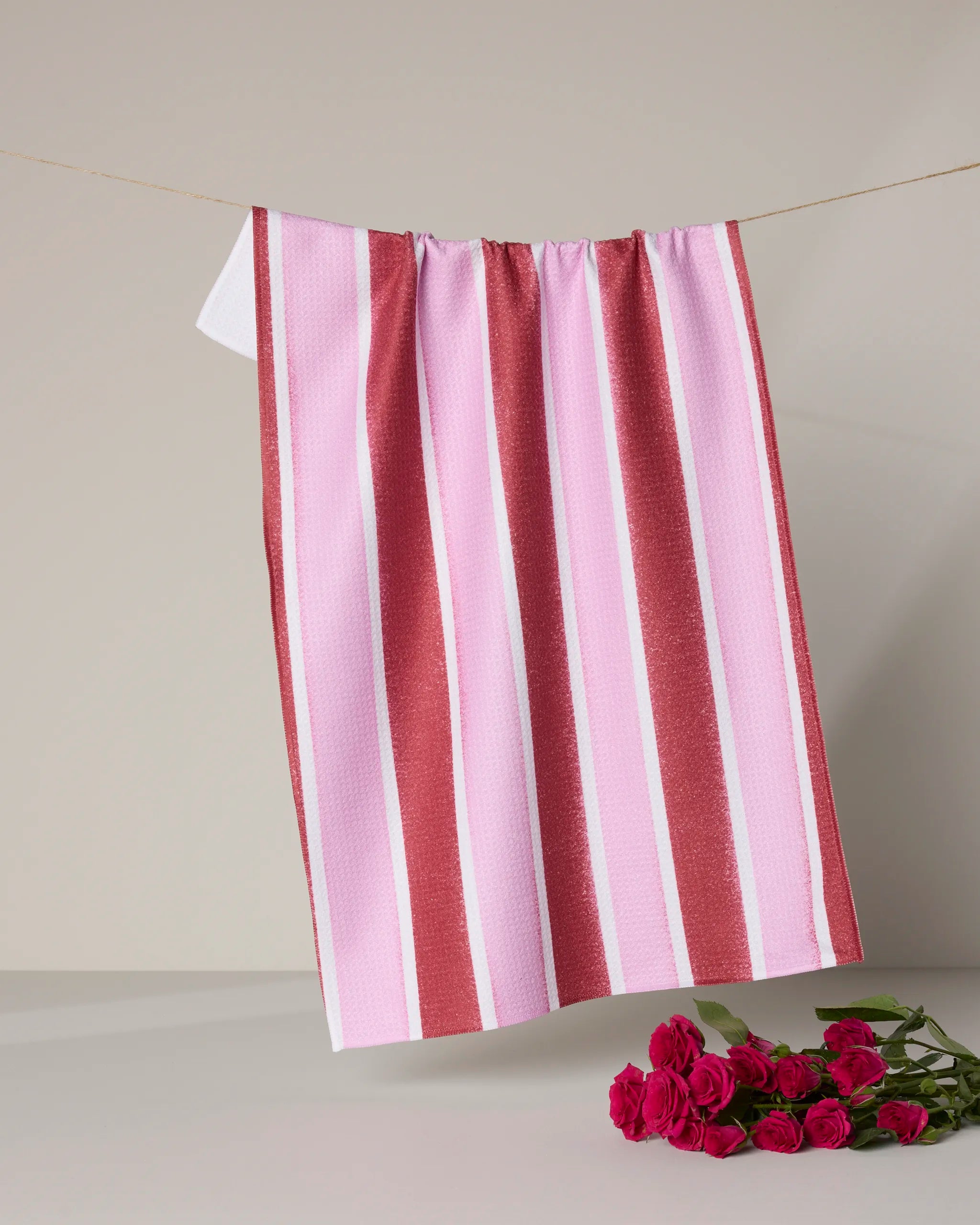 The Geometry Super Sweet Stripe tea towel, featuring pink and white stripes and ultra-absorbent fabric, hangs on a clothesline above a small bouquet of dark pink roses against a simple background.