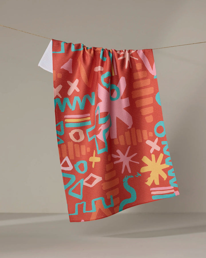 The Sareka Unique "Superstar" towel features playful geometric and squiggle patterns in turquoise, yellow, and pink on a vibrant red-orange base, quick-drying and eye-catching, shown hanging against a plain background.