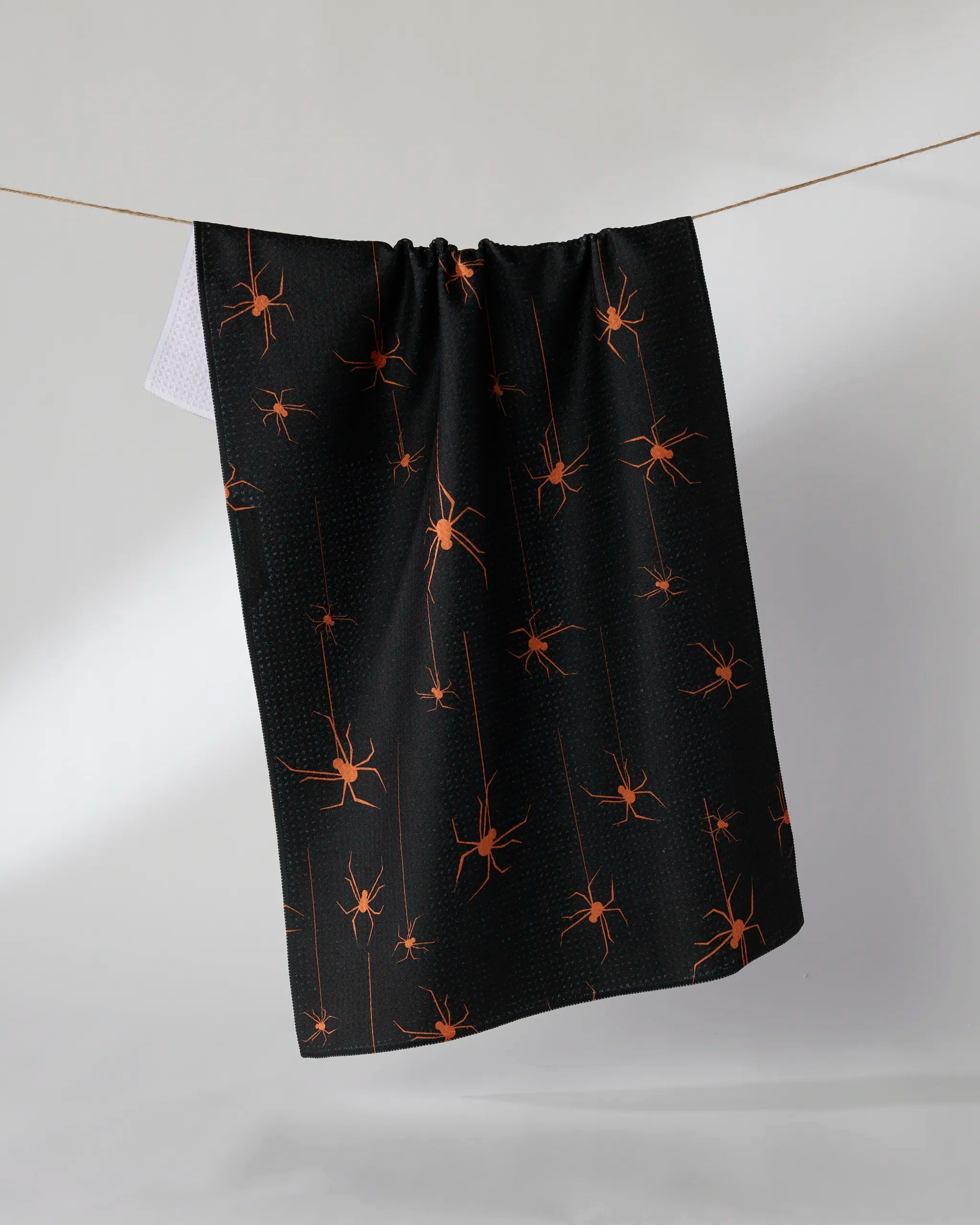 The Suspended tea towel by Geometry hangs on a line, showcasing a repeating pattern of orange spiders and webs on a black background.