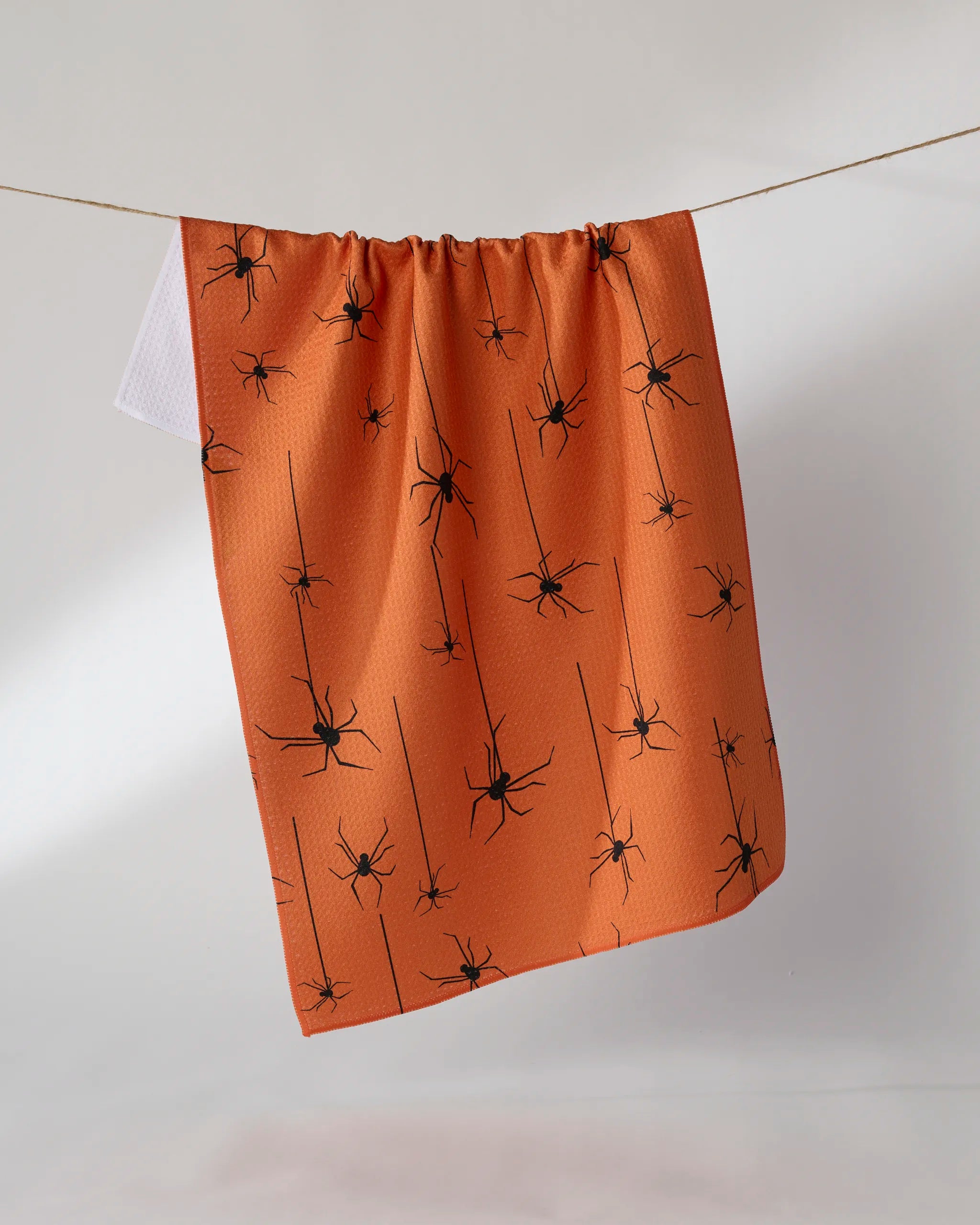 A Geometry "Suspended" towel featuring a black spider pattern in orange quick-drying fabric hangs on a clothesline against a plain white background.