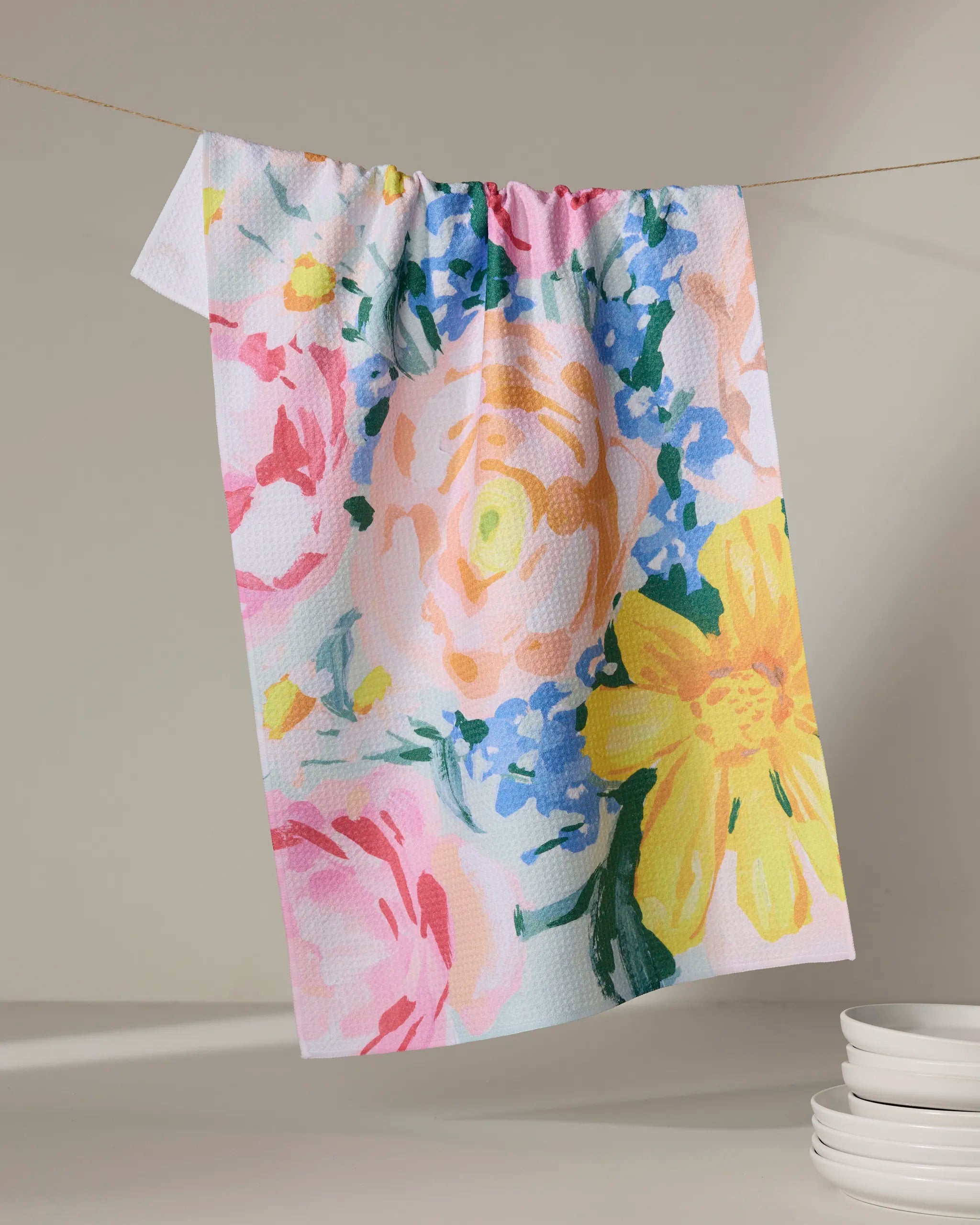 The Suzzy tea towel by Ania Zwara Art features a textured white fabric with a colorful floral print in pink, yellow, blue, and green. This absorbent towel adds charm when displayed near stacked white dishes.