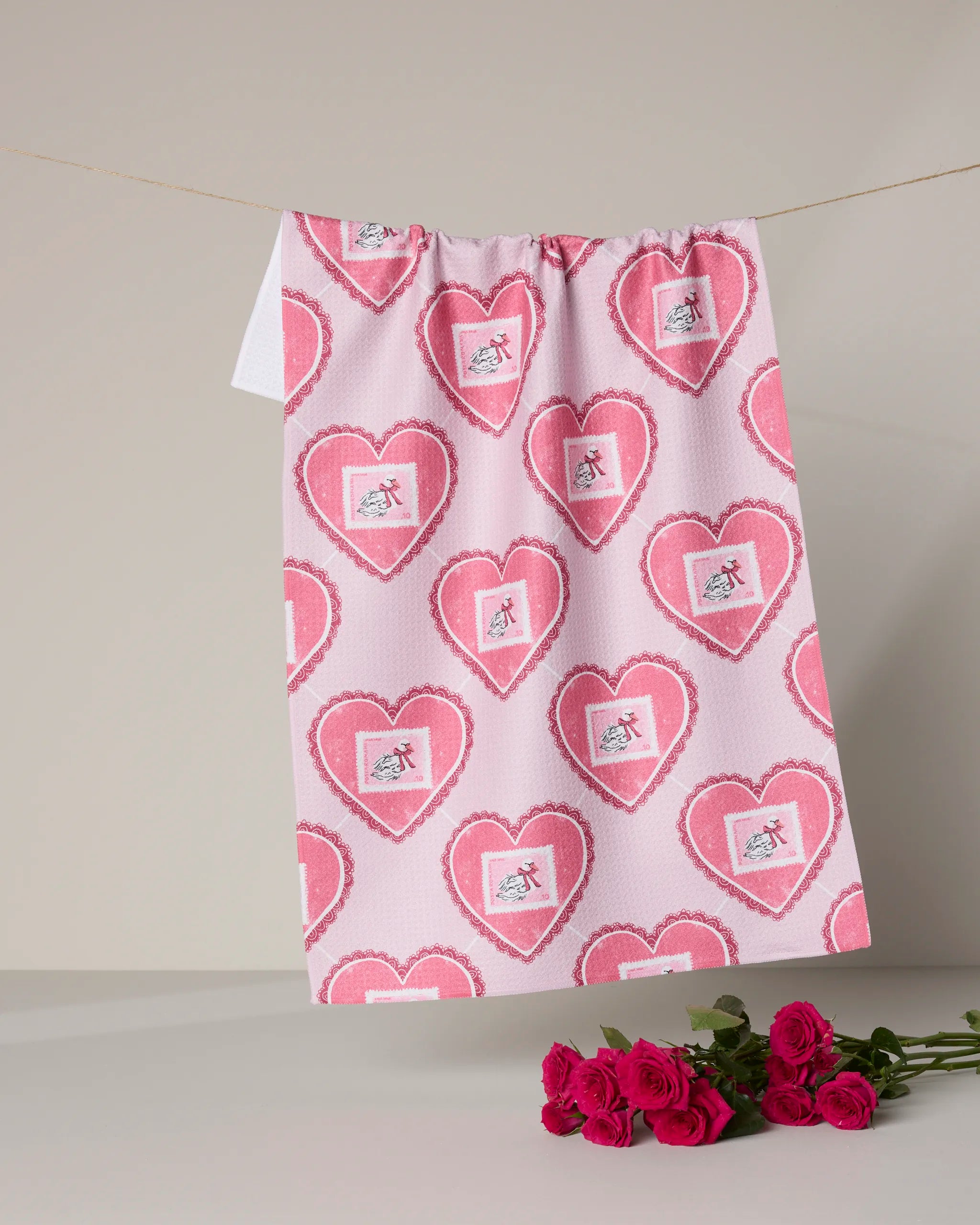 A pink, quick-drying Geometry "Swan Argyle" tea towel with heart and cupid designs hangs on a string against a neutral background, with a bouquet of pink roses placed below.