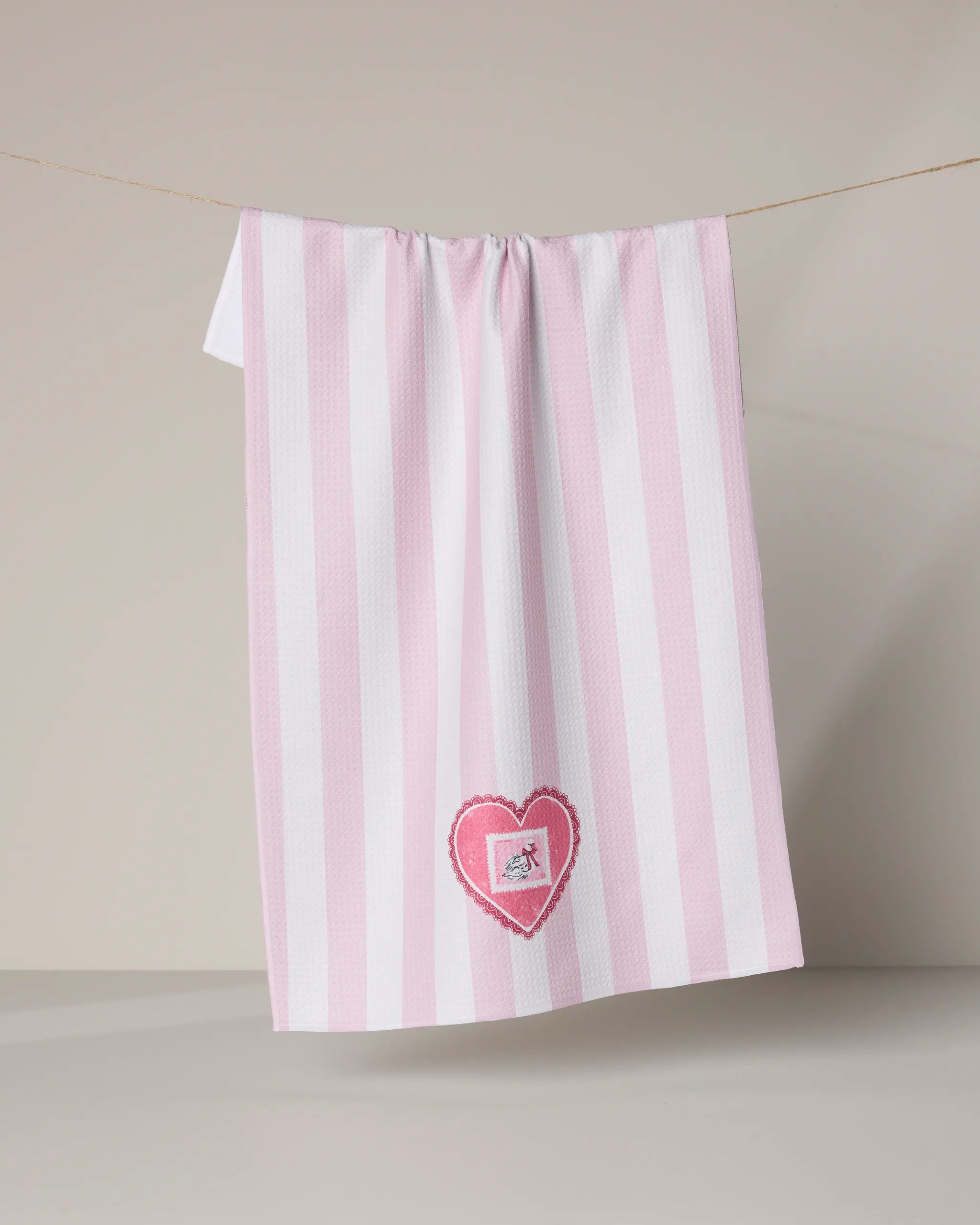 The Geometry “Swan” tea towel features pink and white stripes, a pink heart patch with an embroidered bird near the bottom center, and is made from recycled materials. It hangs on a line against a neutral, minimal background.
