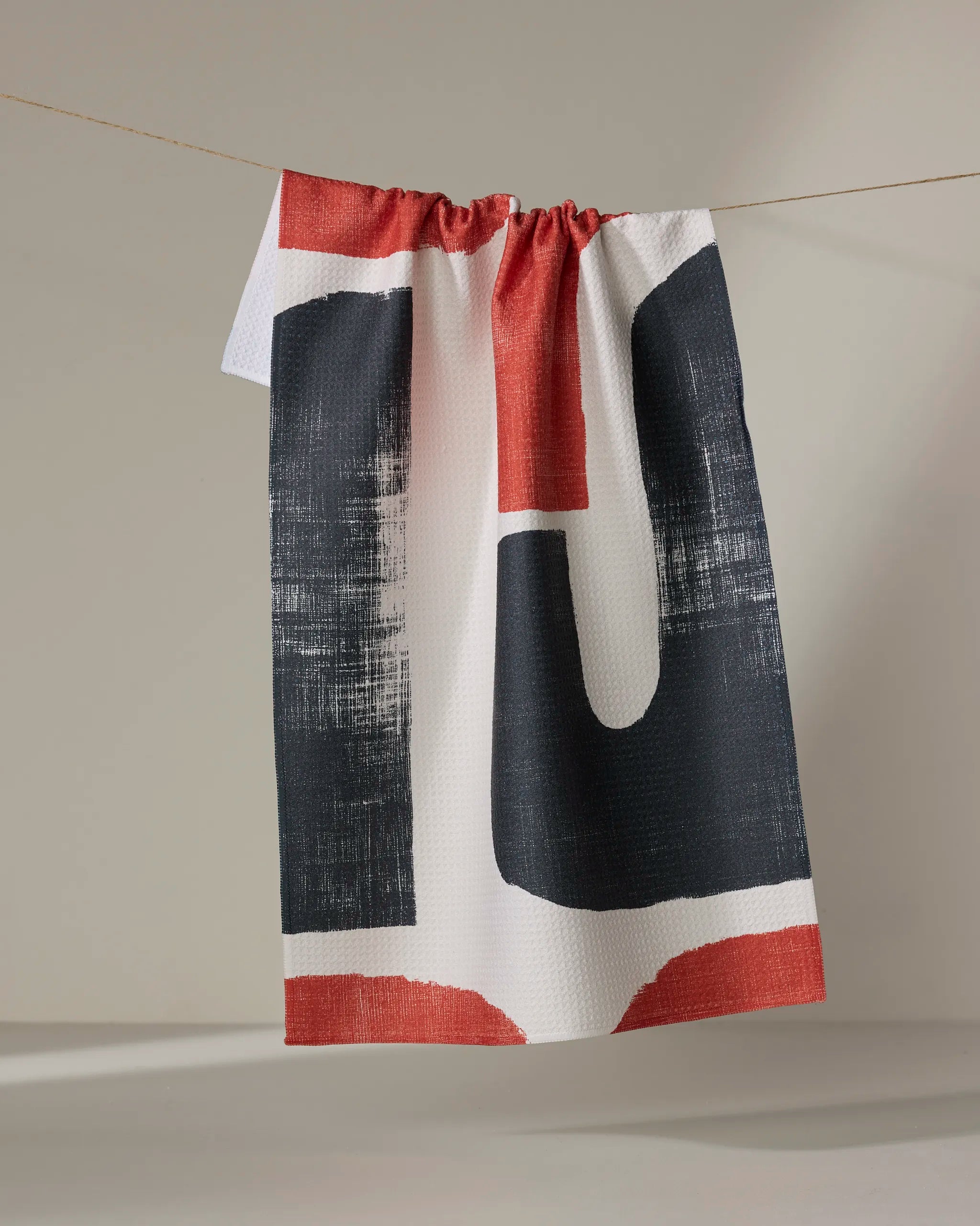 The Sway tea towel by Geometry, featuring a bold black and red abstract design, hangs from a string against a plain, neutral background.