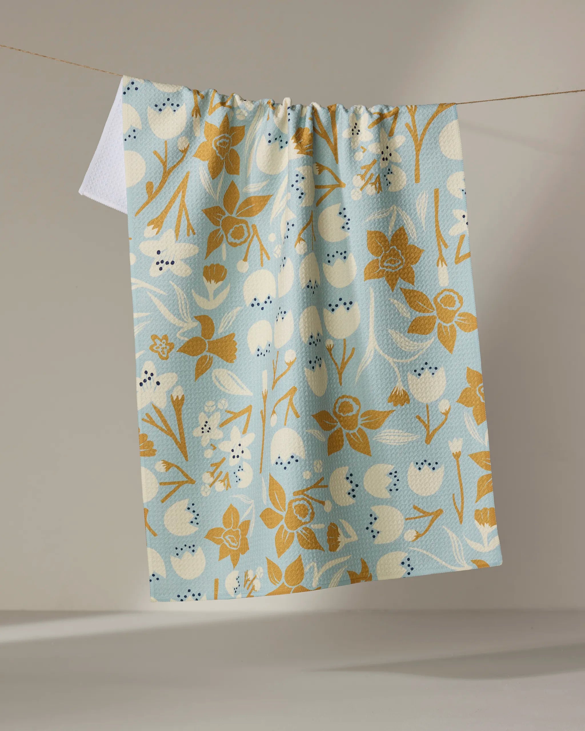 The "Swedish Cherry Blossoms" kitchen towel by Elisabeth Wing features a gold and white floral and berry pattern on light blue, made from recycled materials, hanging on a clothesline against a neutral background.