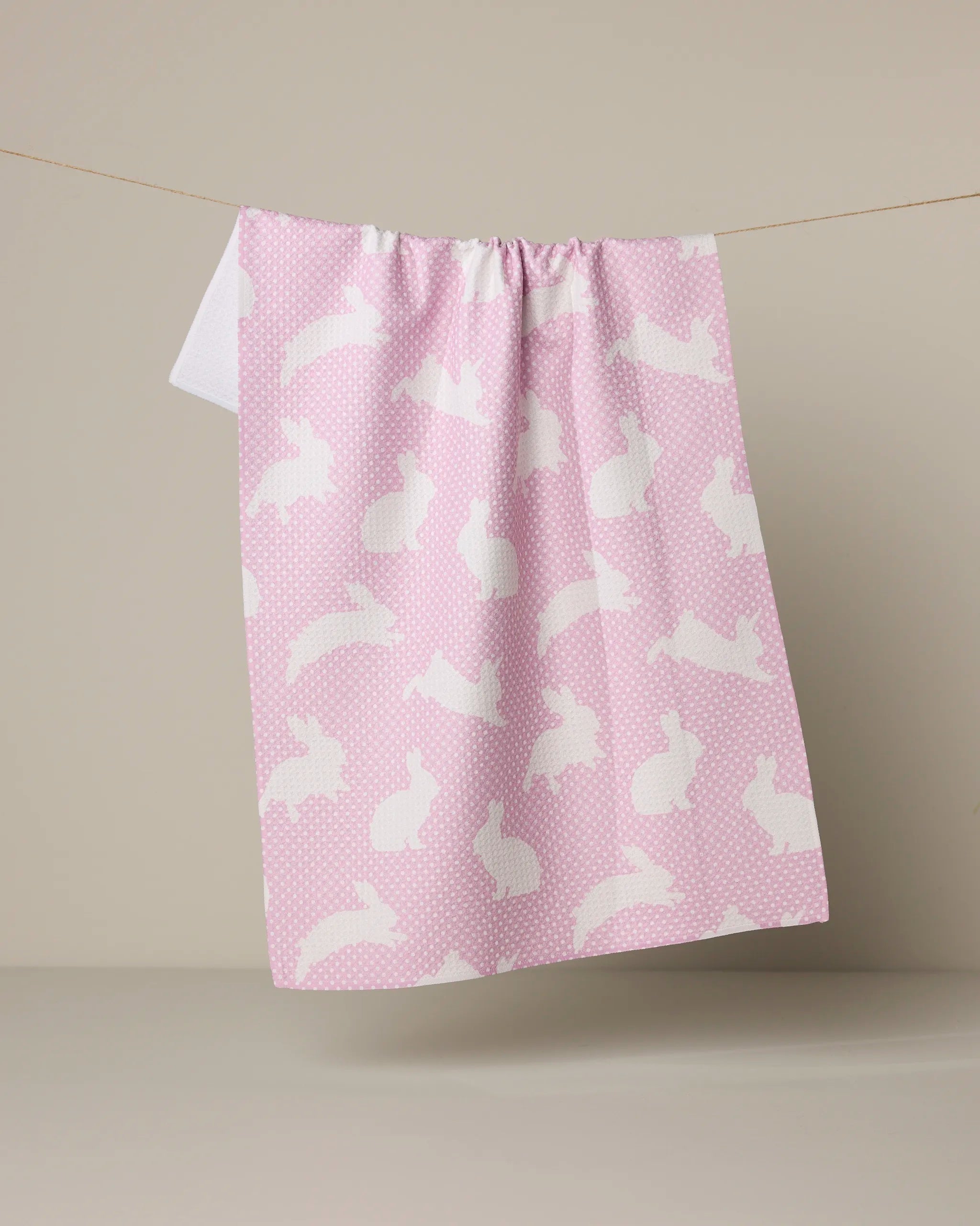 The Geometry Sweet Bunny tea towel, made from recycled materials, features quick-drying pink fabric with white bunny silhouettes and small dots, hanging on a line against a neutral background.