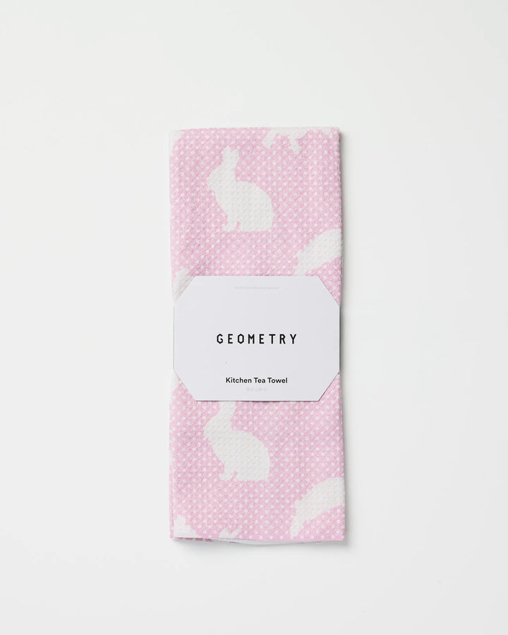 The Geometry Sweet Bunny kitchen tea towel, made from quick-drying recycled materials, features pink fabric with white polka dots and bunny silhouettes. It is folded and wrapped with a GEOMETRY label on a plain white background.