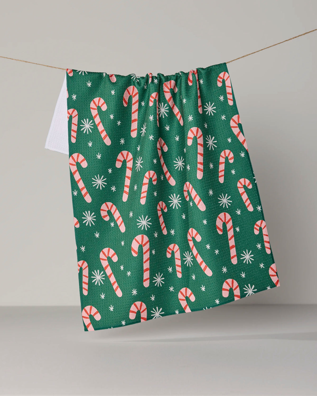 The Geometry Sweet Candy green tea towel, made from recycled materials and featuring a pattern of red and white candy canes with white snowflakes, hangs on a string with a small white towel partially visible behind it.