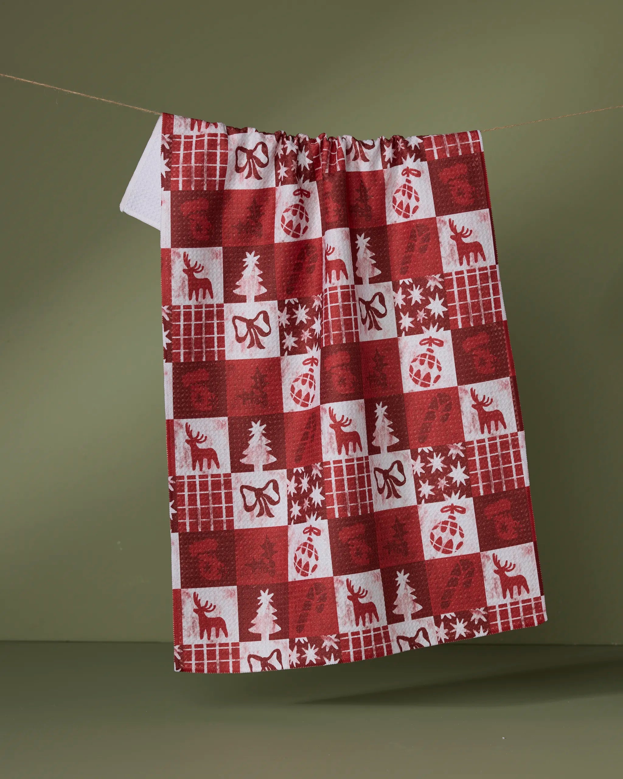 The Geometry Sweet Carol tea towel, featuring a red and white checkered pattern with Christmas motifs like reindeer, bows, snowflakes, pine trees, and ornaments, hangs on a line against an olive green background.