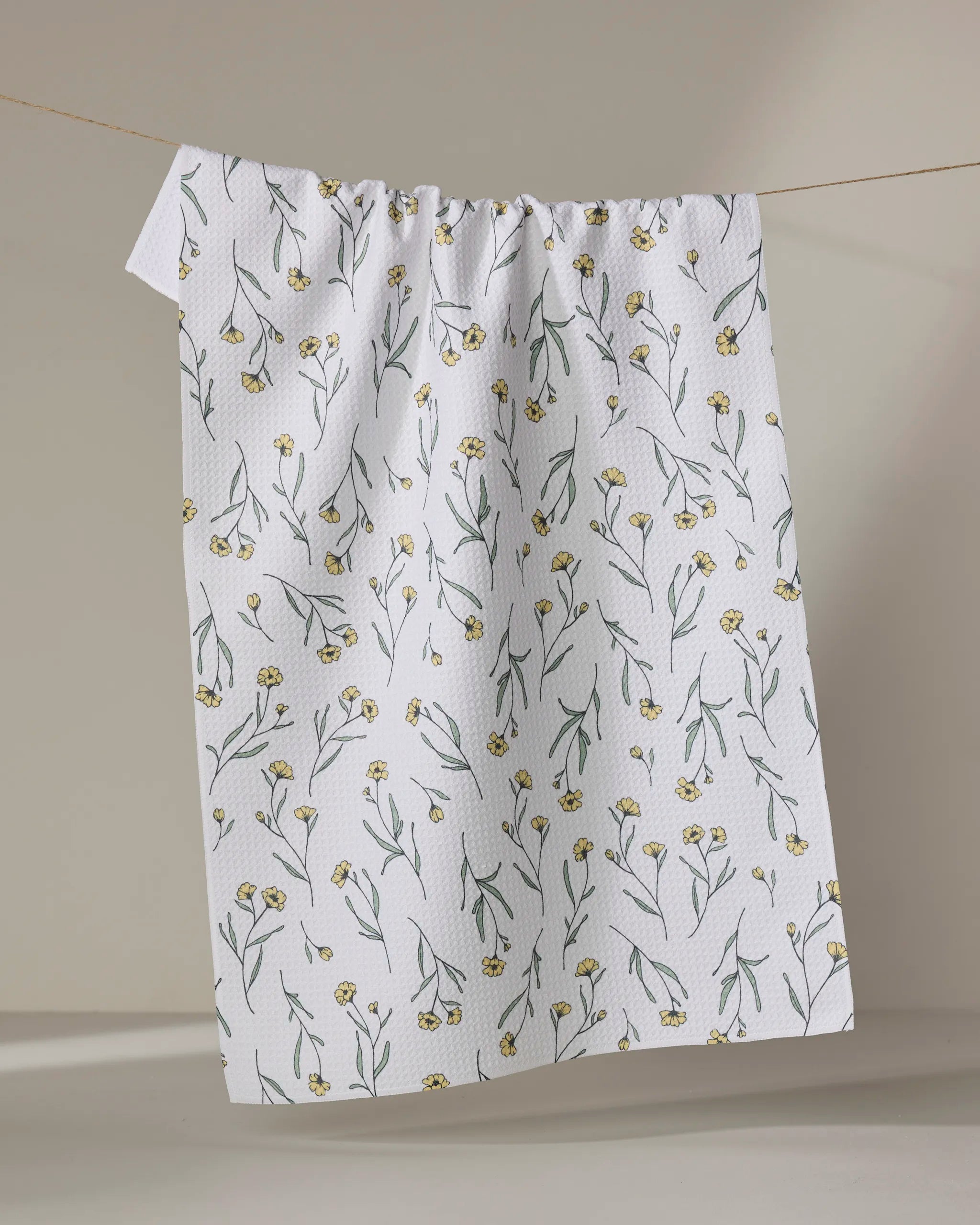 The "Sweet Little Nothings" dish towel by Ashley Celena, featuring yellow flowers with green stems on white, hangs on a clothesline against a light, neutral background.