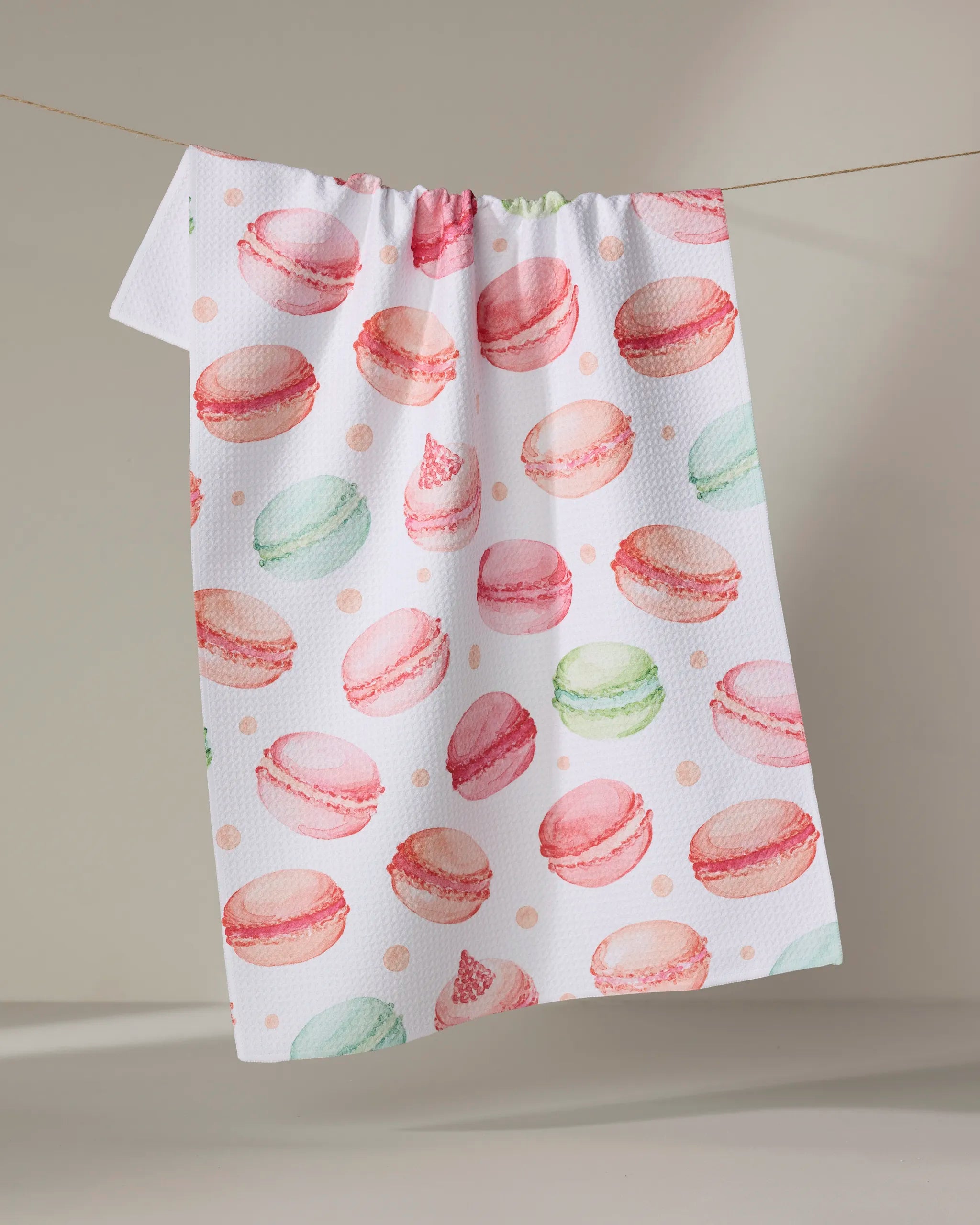 The Elena Fay "Sweet Macarons" white tea towel, crafted from recycled materials and adorned with a vibrant pink, green, and peach macaron print, hangs on a thin string against a neutral background.