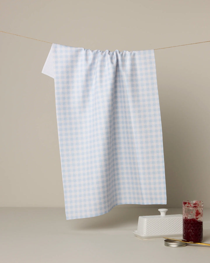 A blue and white checkered Geometry Sweet McKelle kitchen towel hangs from a string against a neutral background, with a butter dish and a jar of red jam displayed below on a light surface.