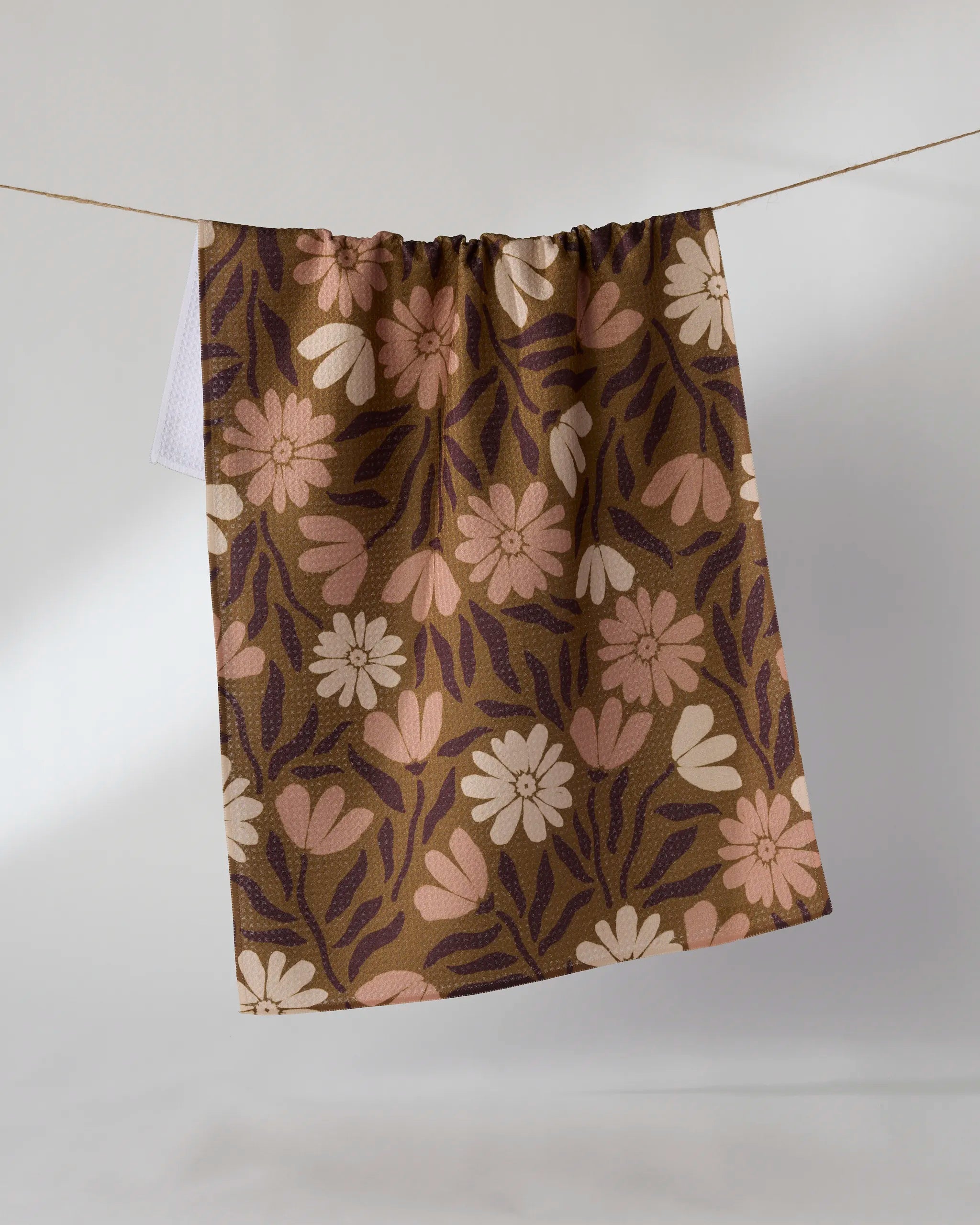 The Geometry Sweet Meadowlane kitchen towel, featuring a cream, pink, and dark purple floral pattern on brown, hangs on a string against a plain light background.
