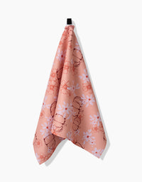 The Sweet Pumpkin Harvest kitchen towel by Ashley Celena hangs from a black loop against a white background, featuring a pink color with floral and beetle patterns and made from recycled materials.