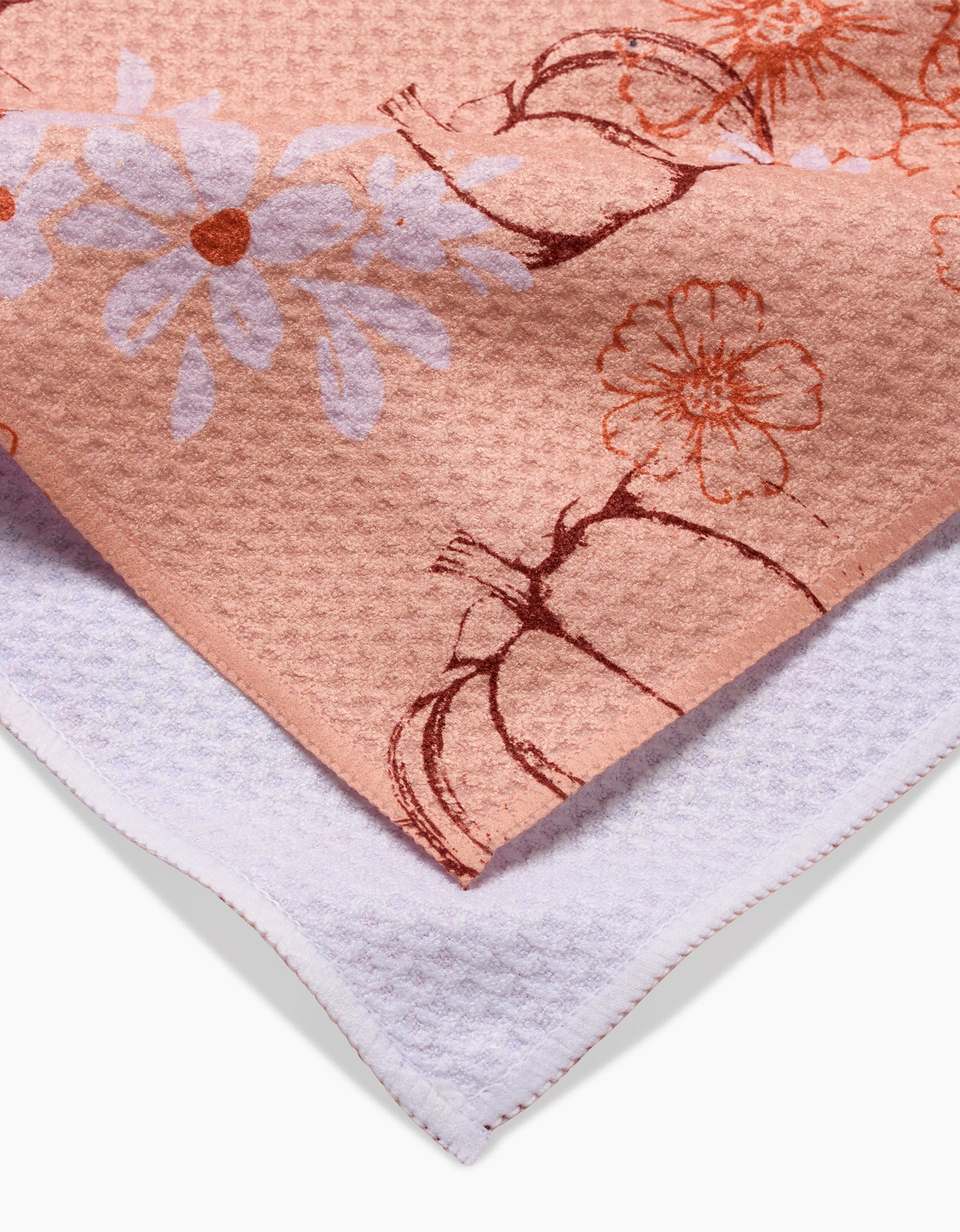 A close-up of two towels: on top is Ashley Celena’s Sweet Pumpkin Harvest tea towel, featuring peach fabric with white and orange floral and pumpkin prints; underneath is a plain, textured white towel.