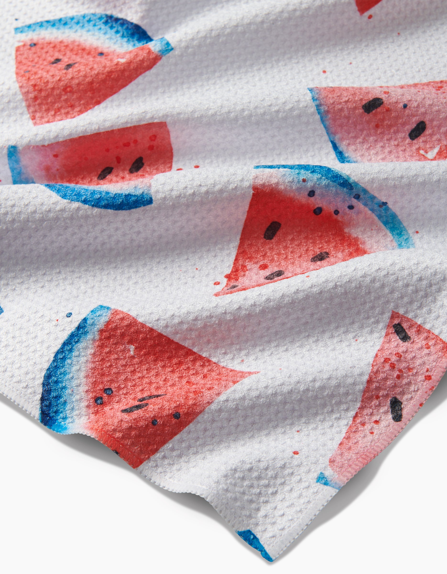 Sweet Slice Of July is a textured white tea towel made from recycled materials, showcasing a vibrant watermelon pattern with red flesh, black seeds, and blue-green rinds.