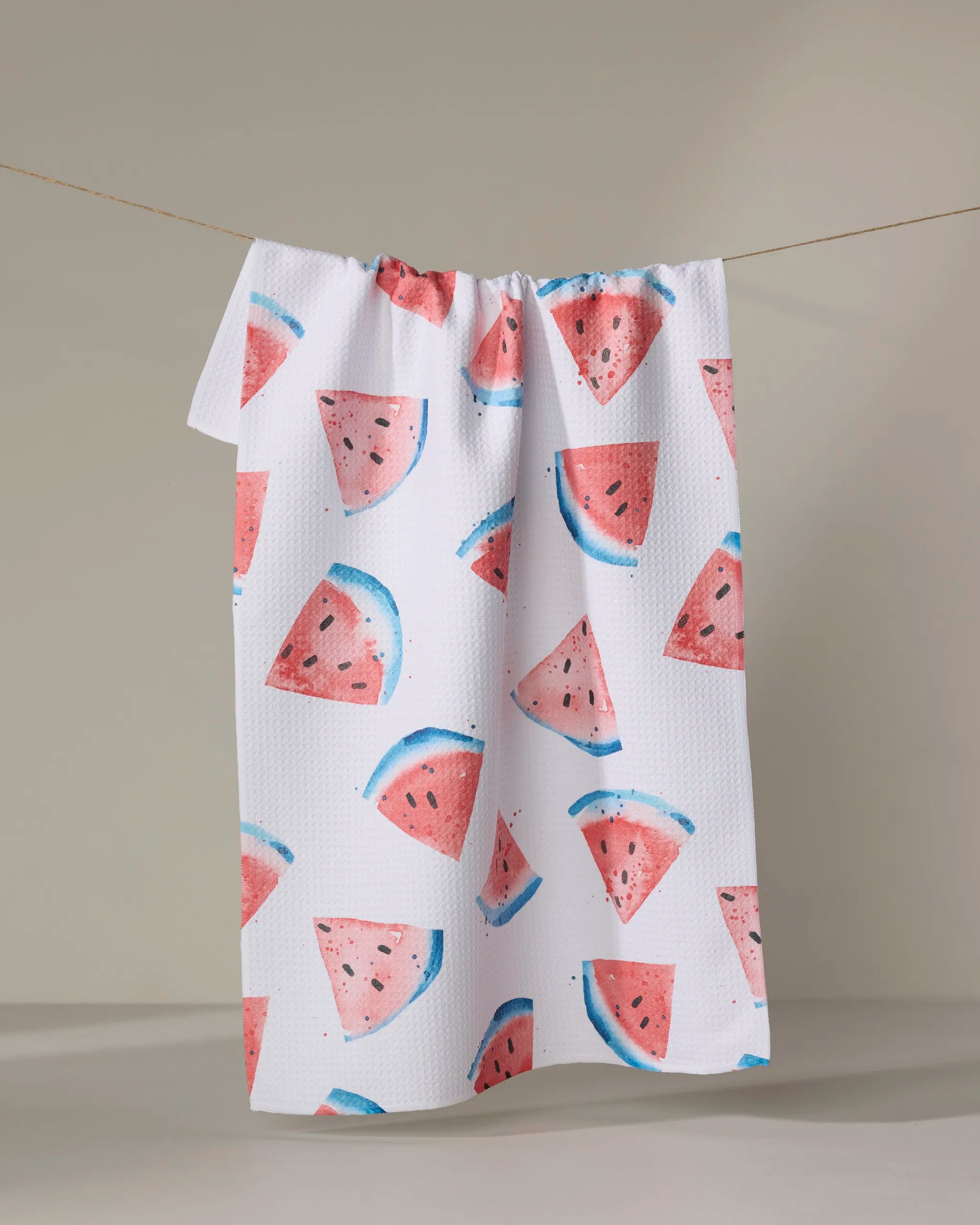 The Sara Mari "Sweet Slice Of July" kitchen tea towel, featuring a pink and blue watermelon slice pattern, hangs on a clothesline against a plain background.