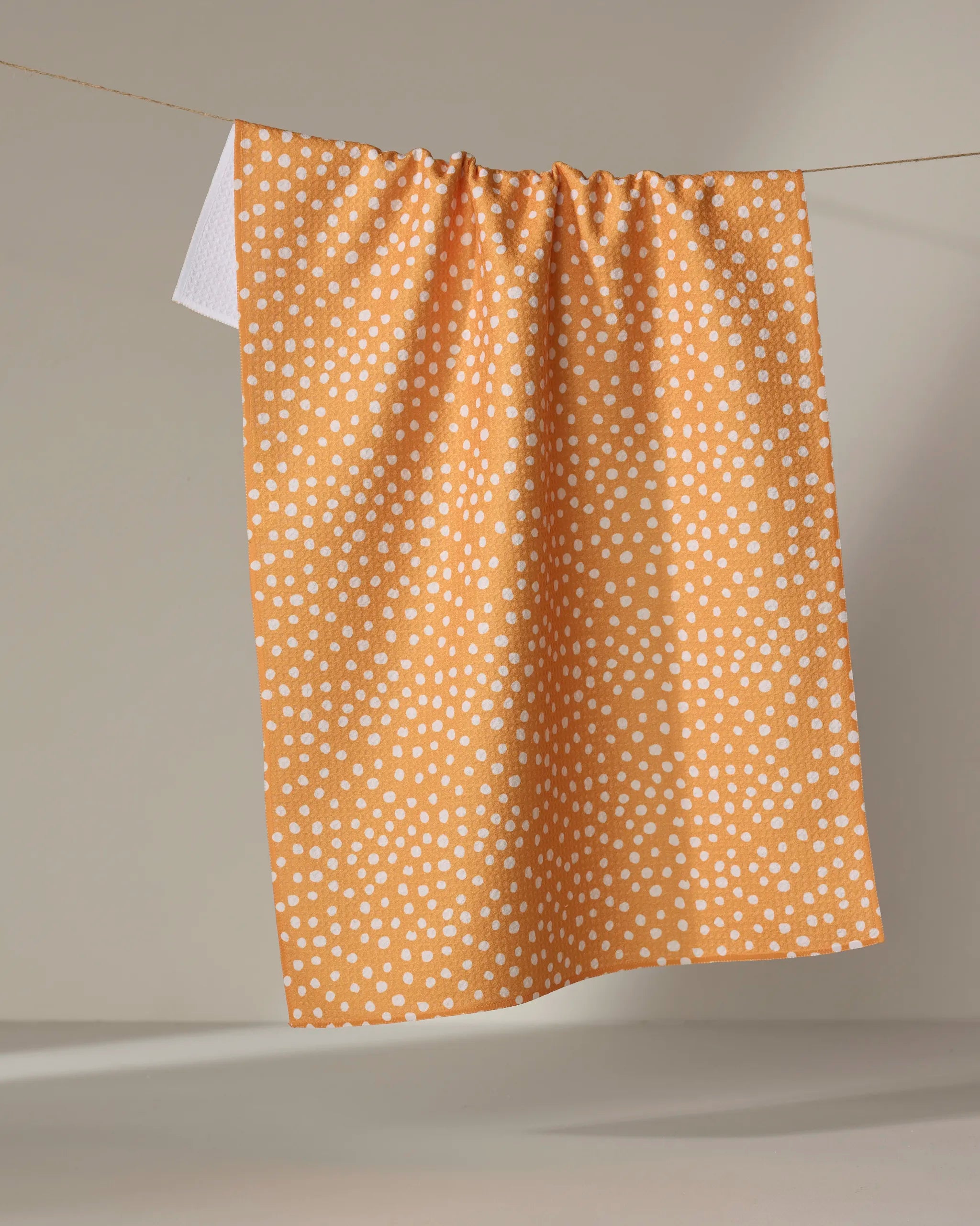 The Sweet Sunshine tea towel by Purely Kaylie, an eco-friendly rectangular orange towel with white polka dots, hangs on a clothesline against a neutral background.