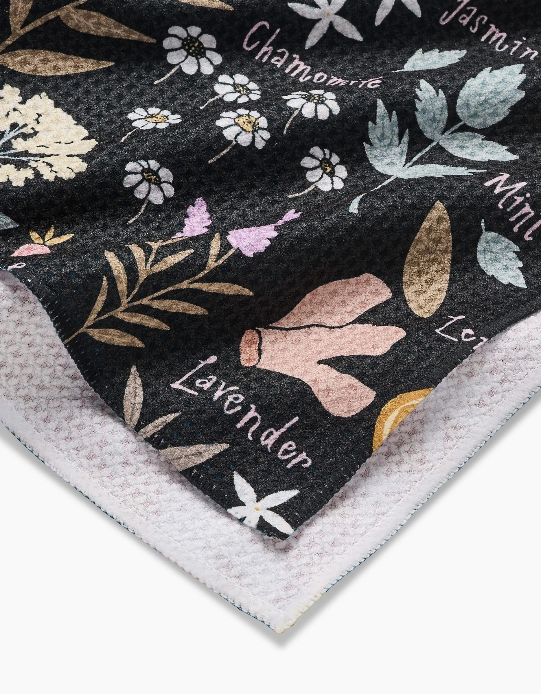 A close-up of the Sweet Tea Geoweave™ tea towel by Janna Sue Design, featuring a soft, eco-friendly fabric with botanical illustrations and labeled herbs like lavender, chamomile, jasmine, and mint on a dark background. One corner is folded.