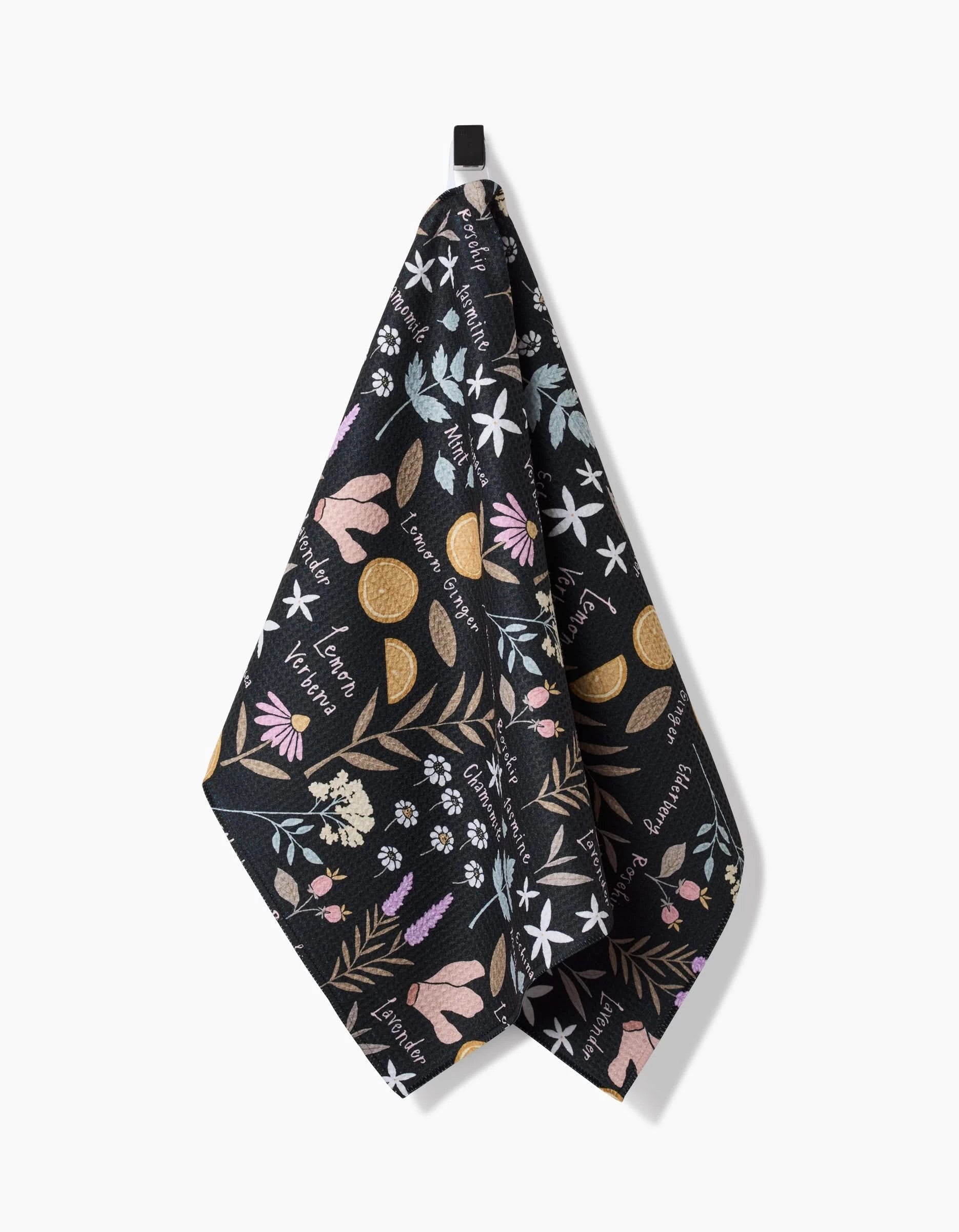 The Sweet Tea tea towel by Janna Sue Design is black, made from recycled materials, and showcases vibrant floral and citrus fruit art with cursive herb names. It hangs from a black loop on a white background.