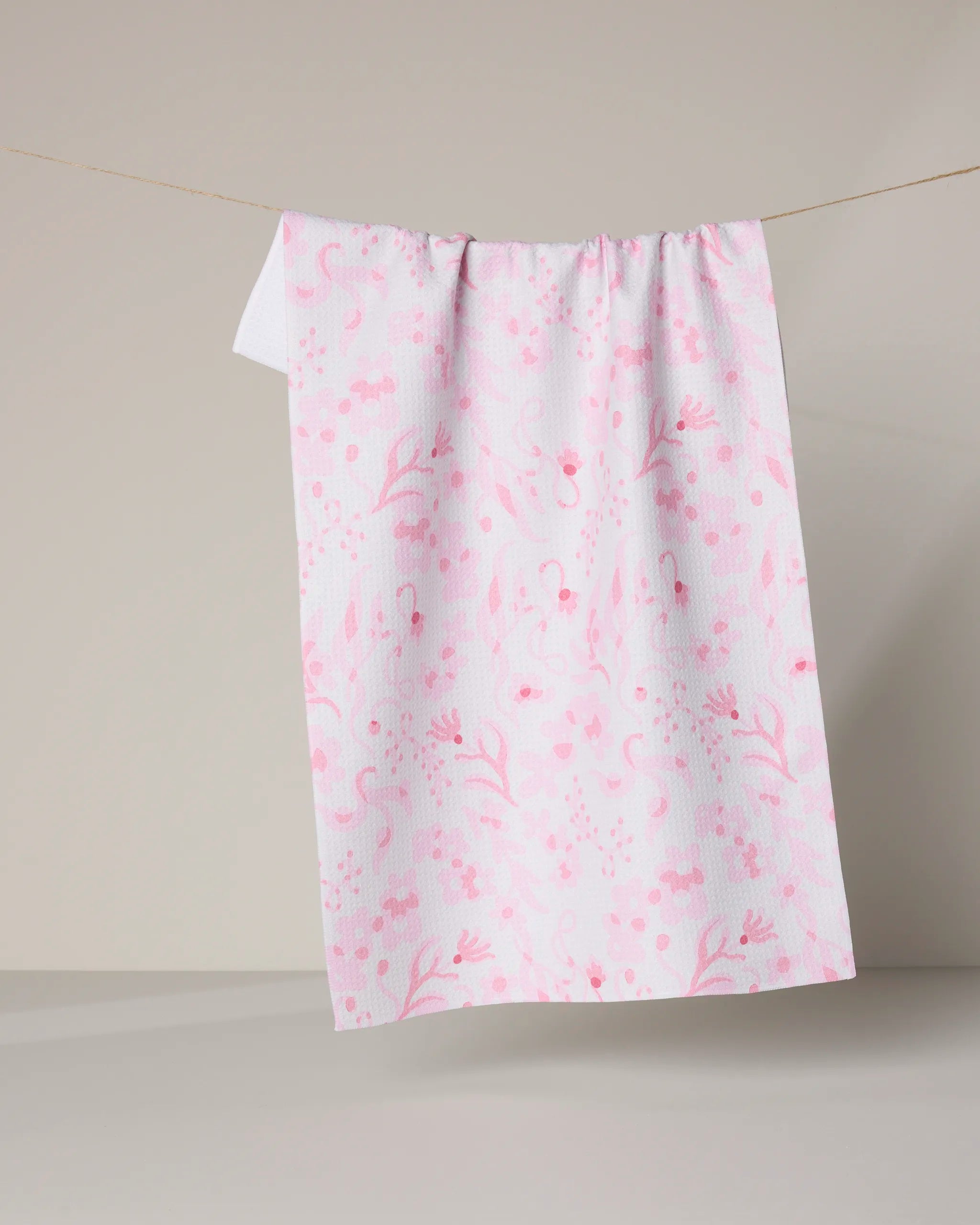 The Geometry "Sweets and Florals" tea towel, made from recycled materials, features a light pink floral and dot pattern on white and hangs on a string against a neutral beige background.