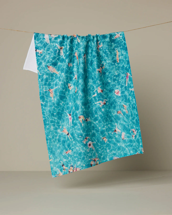 The Geometry "Swimming Crowd" tea towel, made from recycled materials, showcases swimmers in a blue pool and hangs on a clothesline against a plain beige background.