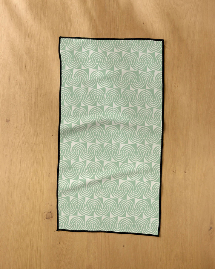 The Geometry Swirl Land fitness towel, featuring a light green and white geometric swirl pattern with black edges, is ultra-absorbent and perfect for active lifestyles. It’s shown laid flat on a light wooden surface.