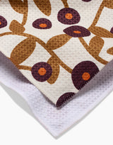 A close-up of Lizzie Clarks Swirling Vines tea towel shows its quick-drying fabric with brown stems, purple flowers with orange centers on a white background, and the folded design reveals both the patterned front and plain white back.