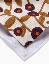 A close-up of Lizzie Clarks Swirling Vines tea towel shows its quick-drying fabric with brown stems, purple flowers with orange centers on a white background, and the folded design reveals both the patterned front and plain white back.