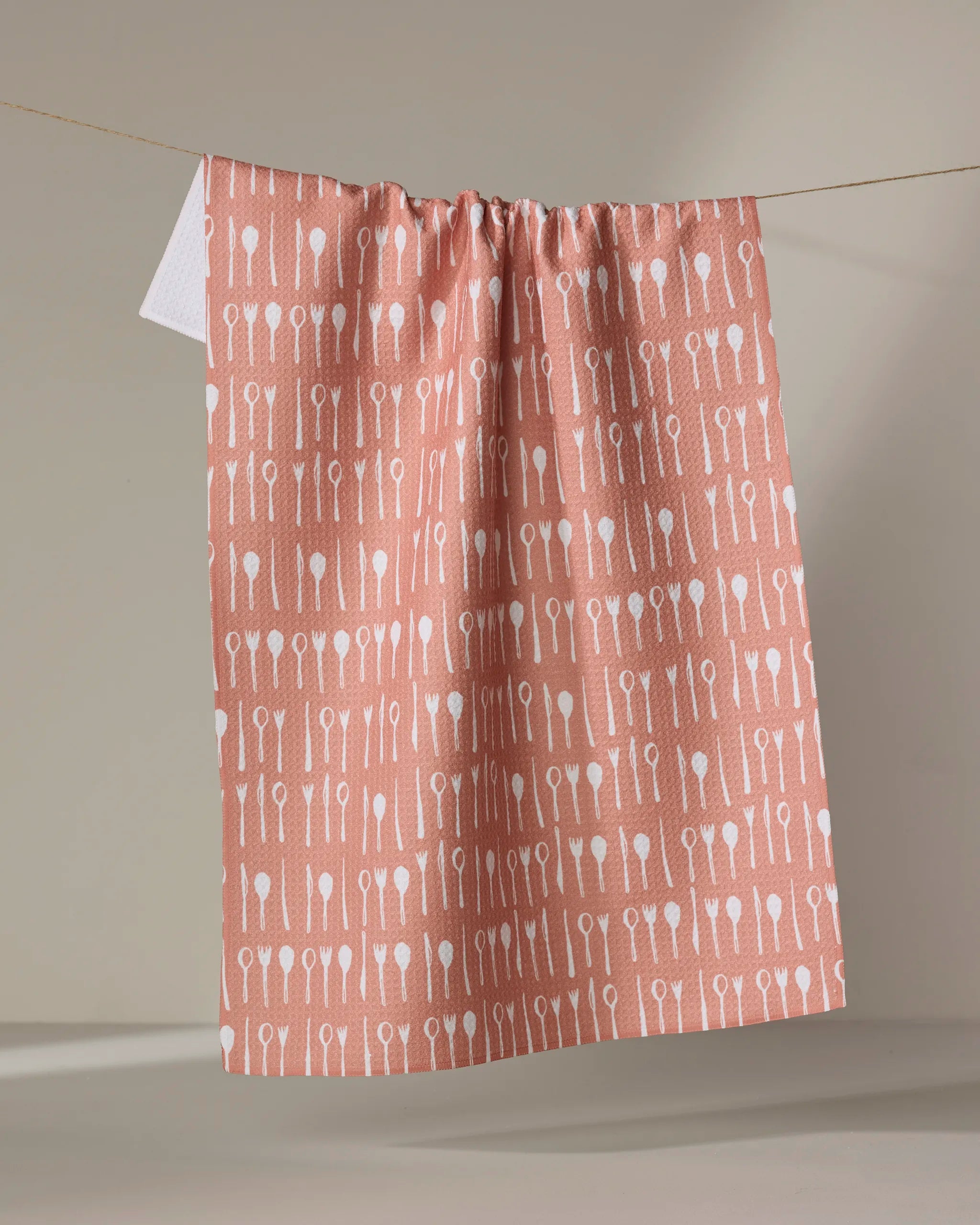 The "Seventies Retro Flowers" tea towel by Claire Iglesias, in pink with a white retro flower pattern, hangs on a string against a neutral background.