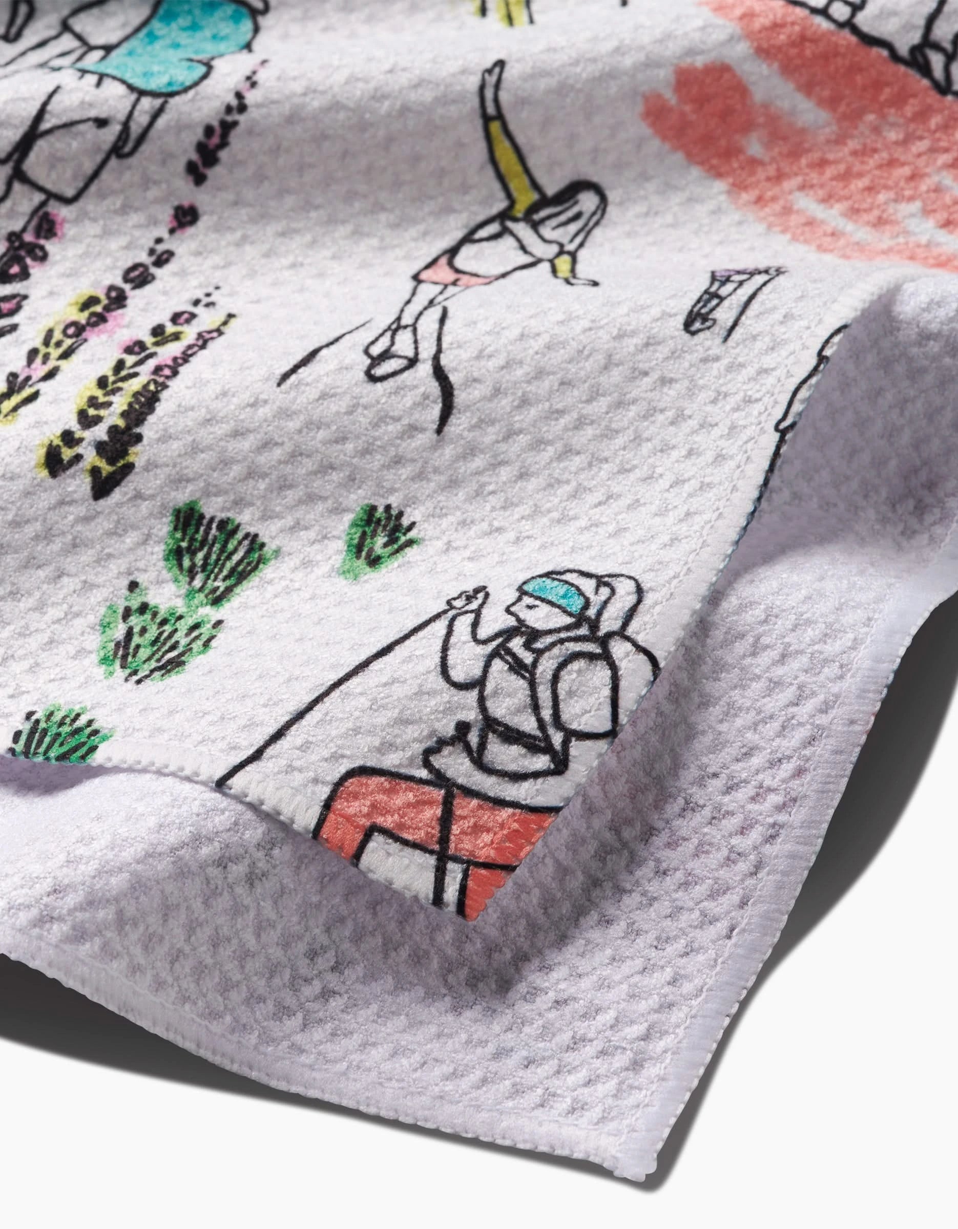 Close-up of the Take A Hike tea towel by Elisabeth Wing, crafted from recycled materials and featuring vibrant, hand-drawn hikers, greenery, and abstract shapes. The textured white fabric is partly folded to display the detailed artwork.