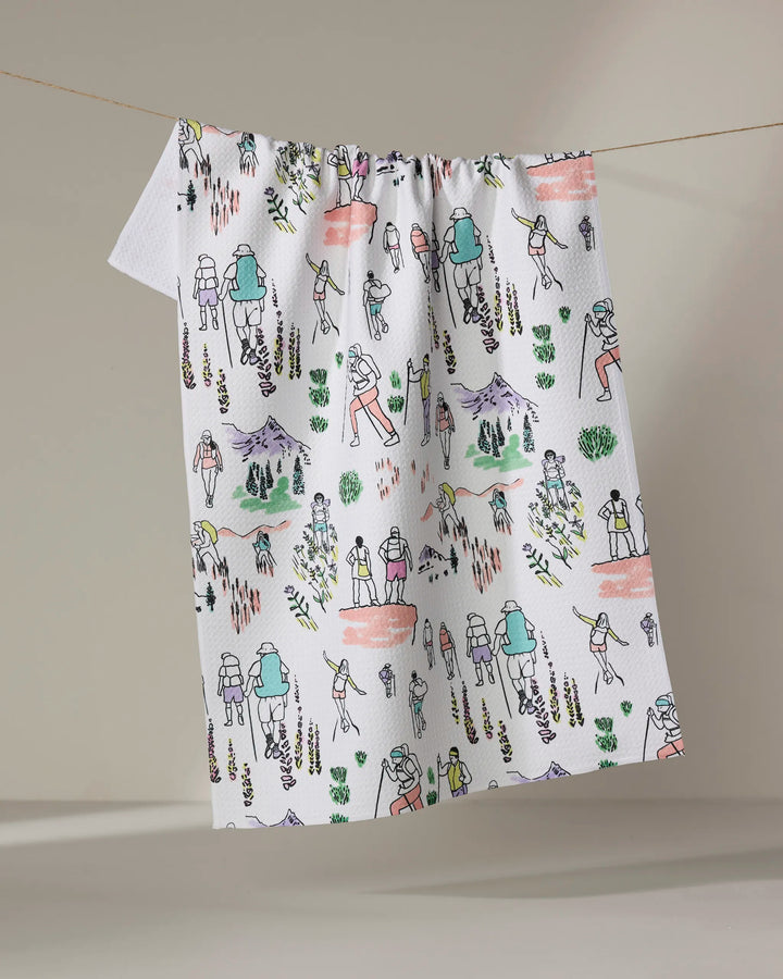 The "Take A Hike" tea towel by Elisabeth Wing features colorful, hand-drawn style illustrations of hikers, mountains, trees, and nature scenes in green, orange, purple, and yellow on white.