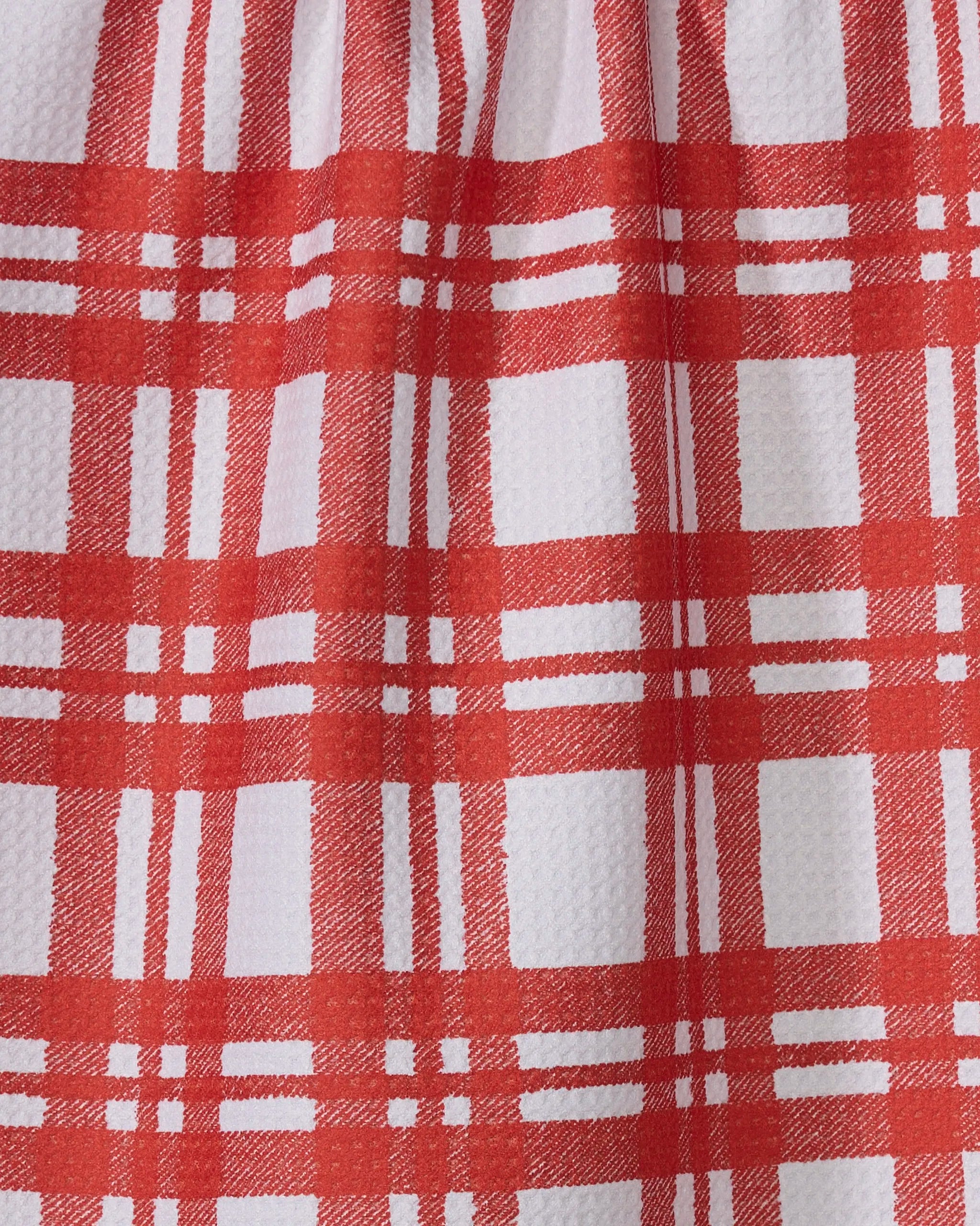 A close-up of Geometry’s eco-friendly tea towel, Tartan, in white with a red plaid pattern of intersecting lines. The fabric’s texture and gentle folds are visible.