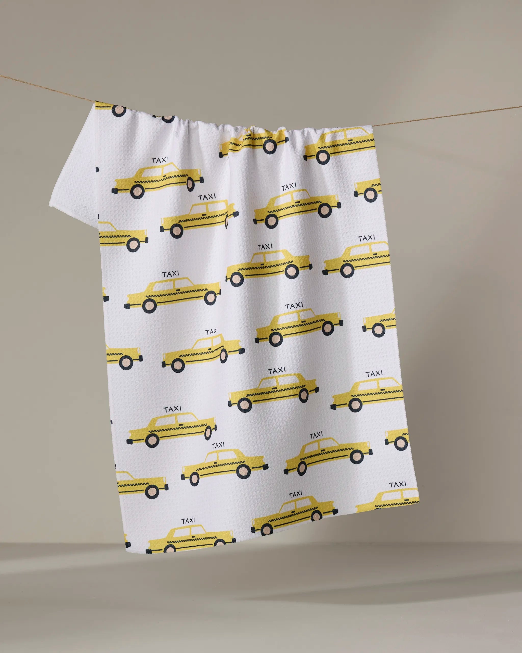 The "Taxi Cabs" tea towel by Vaishali Design Studio features a yellow taxi print and hangs on a clothesline against a neutral background.