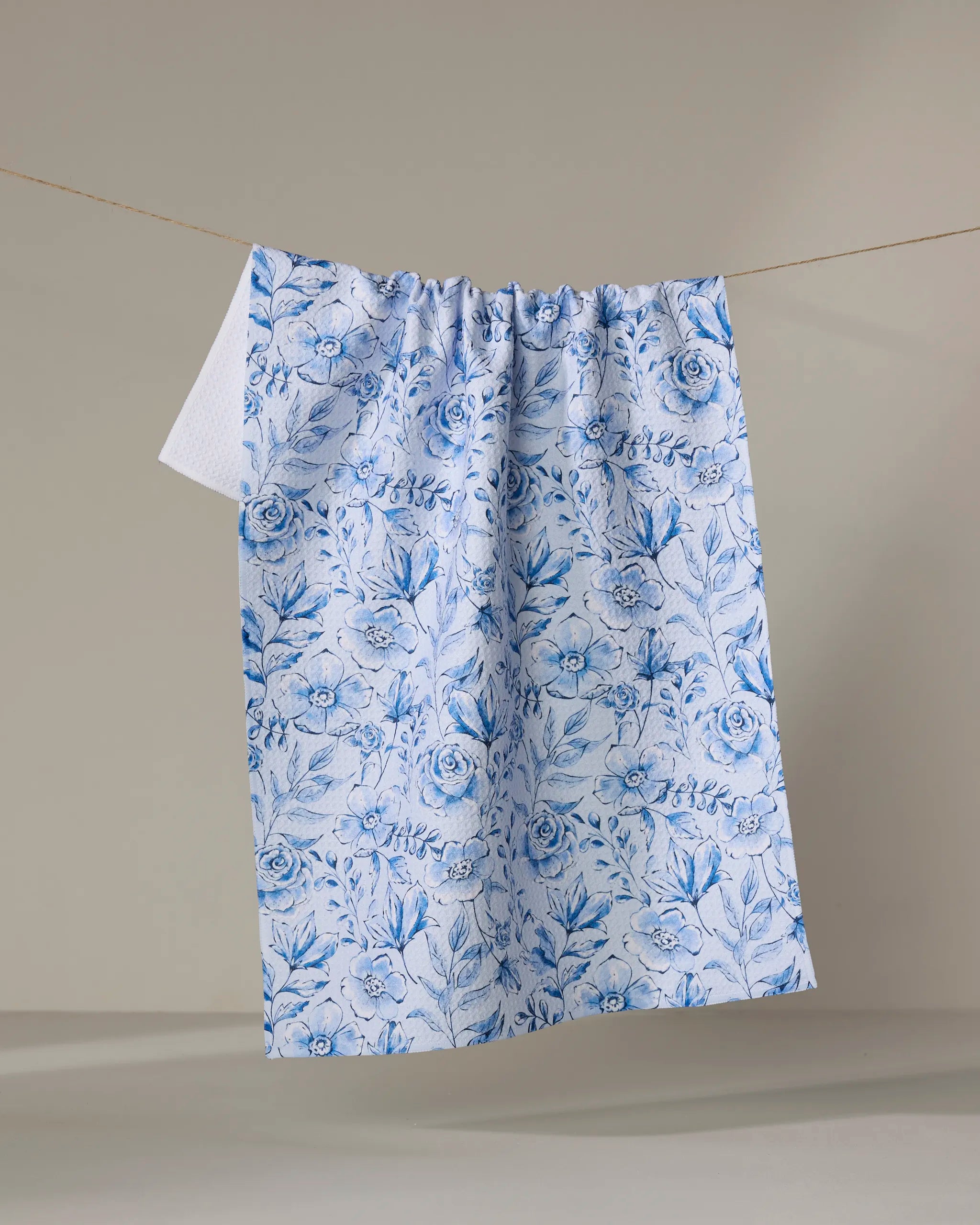 The Sara Mari “Tea Set Blossoms” recycled kitchen towel, adorned with intricate blue and white floral and leaf patterns, hangs on a line against a plain beige background.