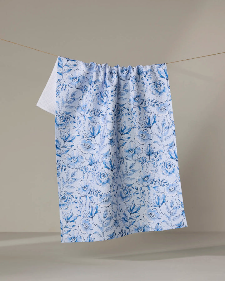The Sara Mari “Tea Set Blossoms” recycled kitchen towel, adorned with intricate blue and white floral and leaf patterns, hangs on a line against a plain beige background.