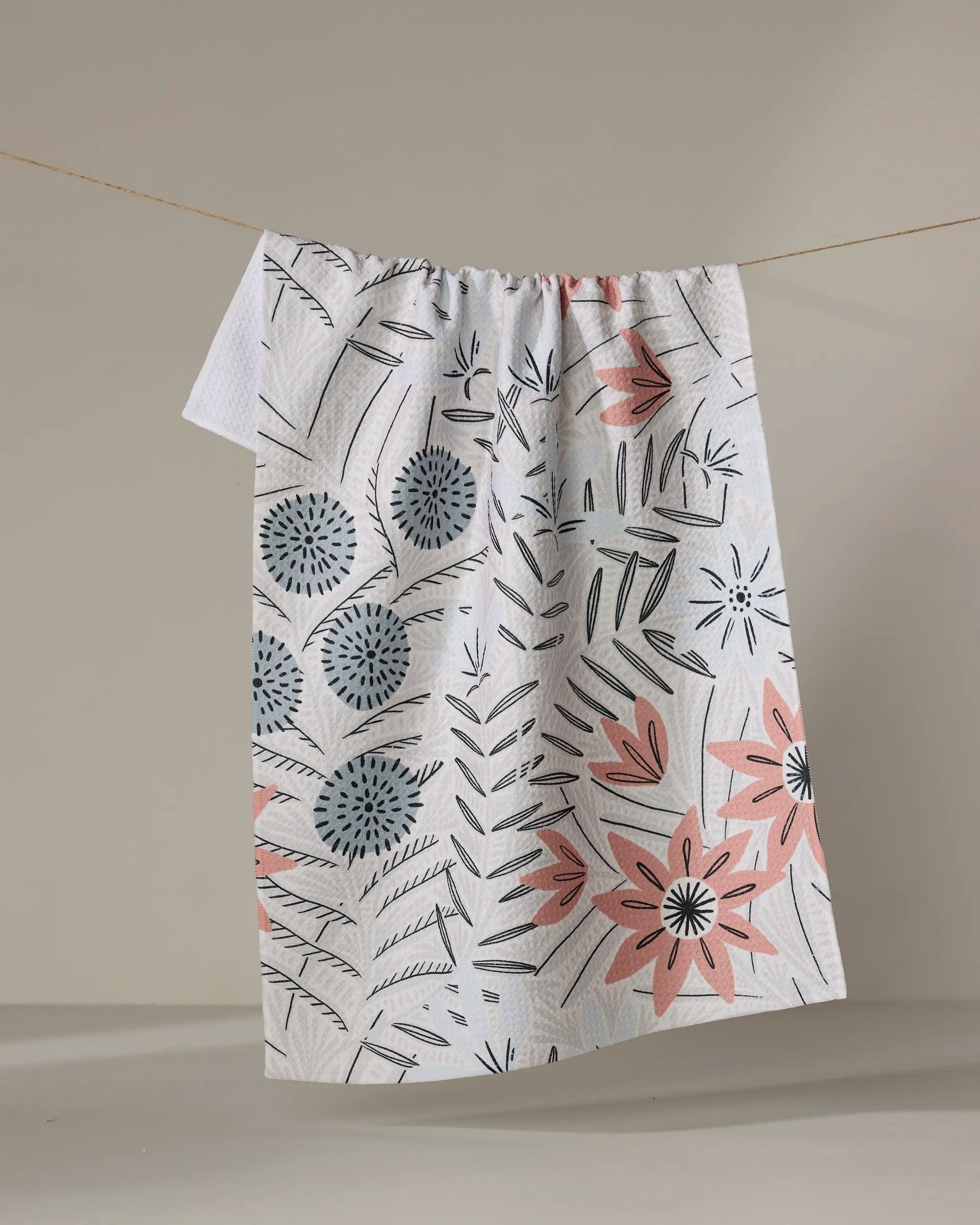 The Teagan Cream dish towel by Amy MacCready, featuring a quick-drying white fabric with soft pink, blue, and gray floral and leaf patterns, hangs on a clothesline against a neutral background.