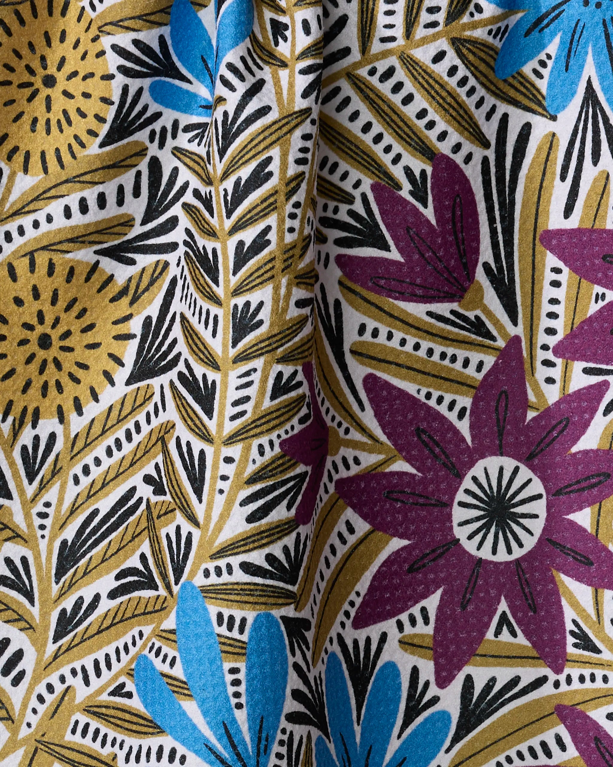 A close-up of the Teagan Dark Maroon tea towel by Amy MacCready shows its colorful floral pattern on a white background, with yellow, purple, and blue flowers, black leaves, and soft fabric wrinkles adding texture and shadows.