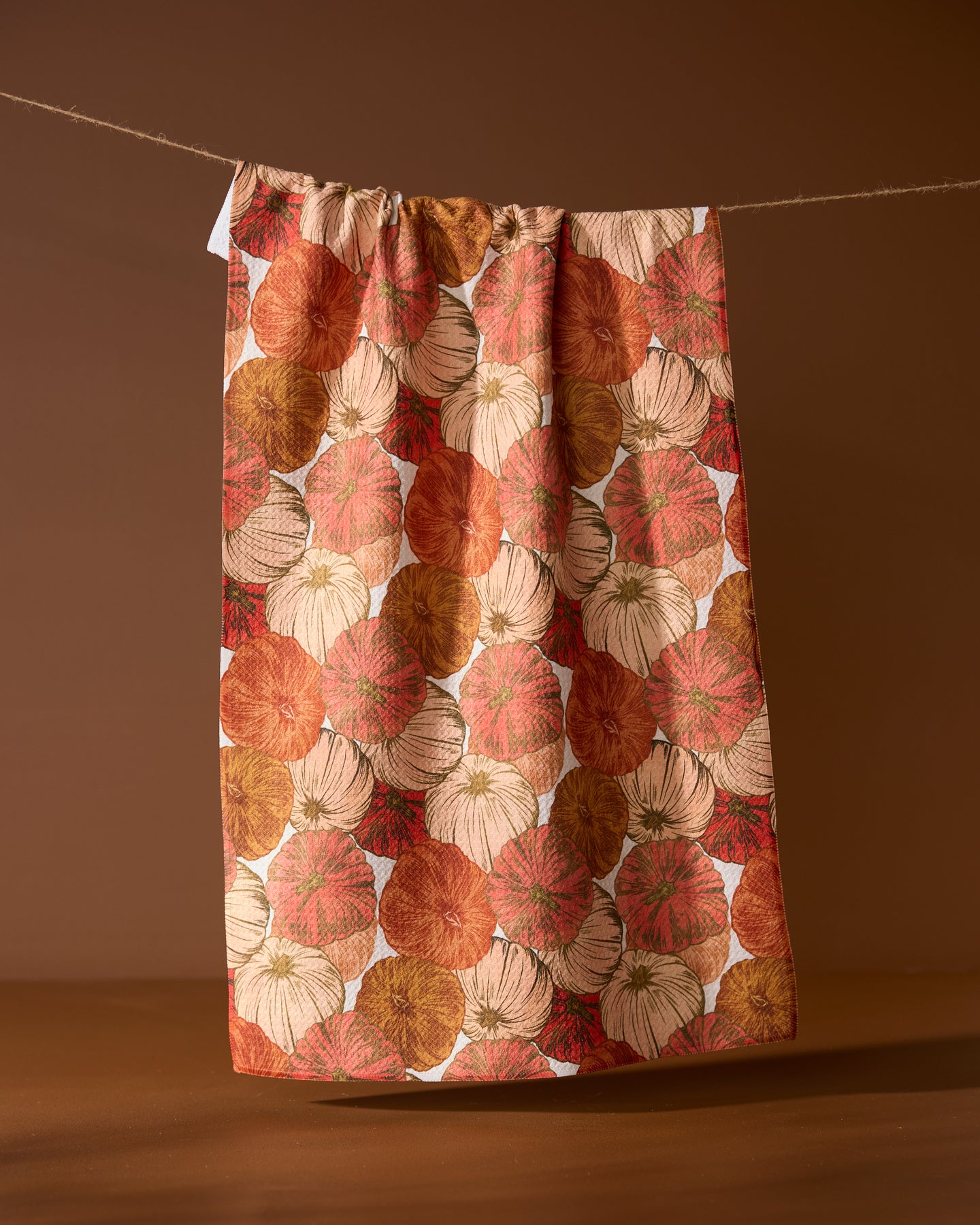 The Geometry Tendril tea towel, featuring absorbent fabric and an autumnal pumpkin pattern in orange, brown, and cream tones, hangs on a clothesline against a brown background.