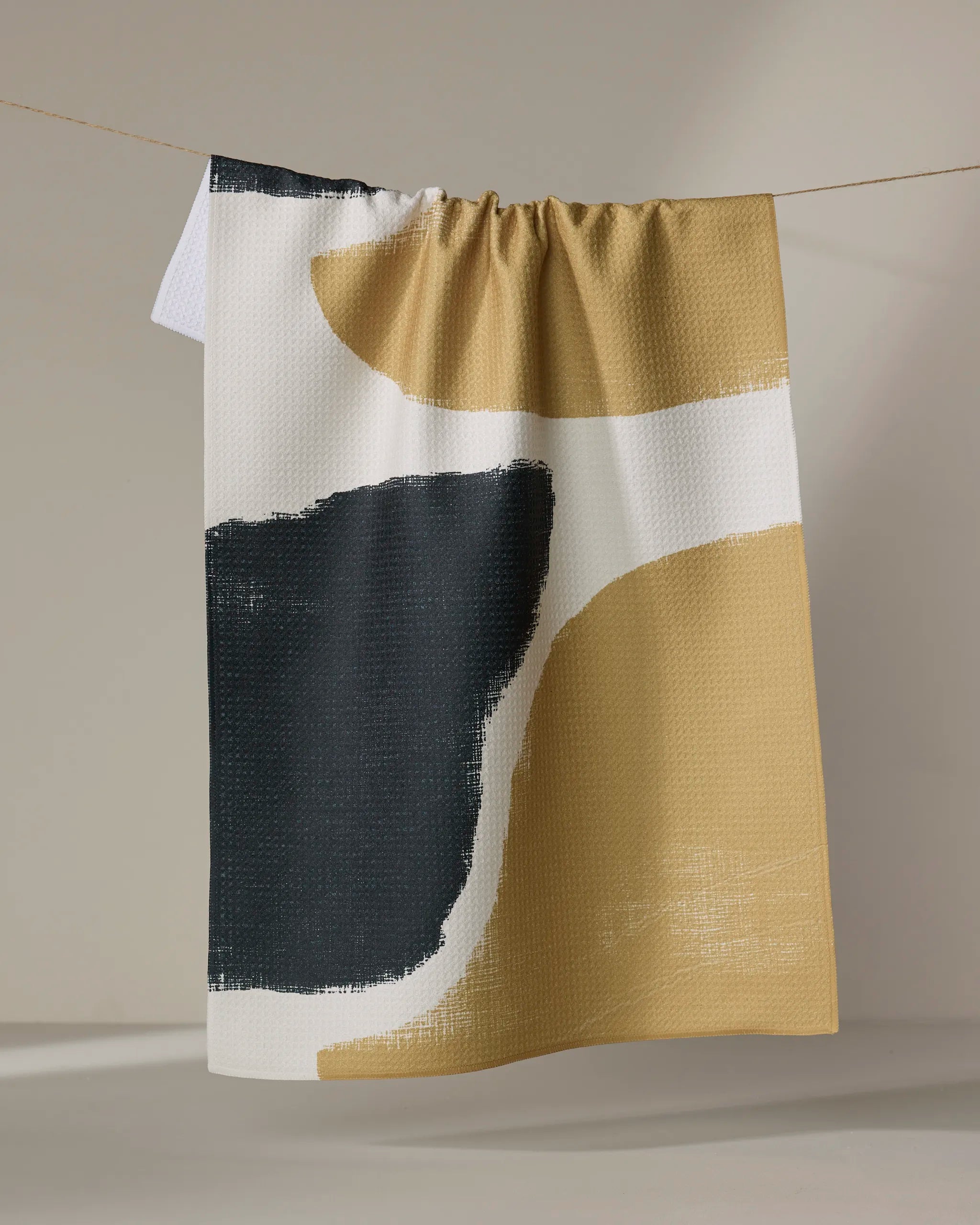 The Terzo kitchen tea towel by Geometry features abstract black, white, and mustard yellow brushstrokes and hangs on a string against a neutral background.