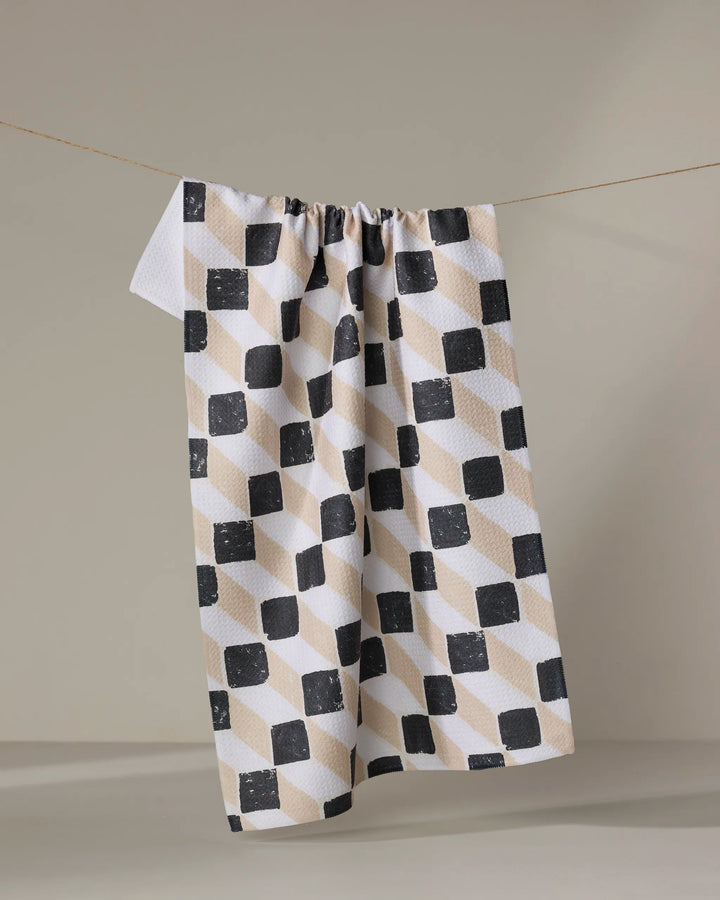 The Tessera kitchen towel by Studio Jeannot, crafted from recycled materials, features a geometric black, beige, and off-white cube pattern and hangs on a string against a plain light background.